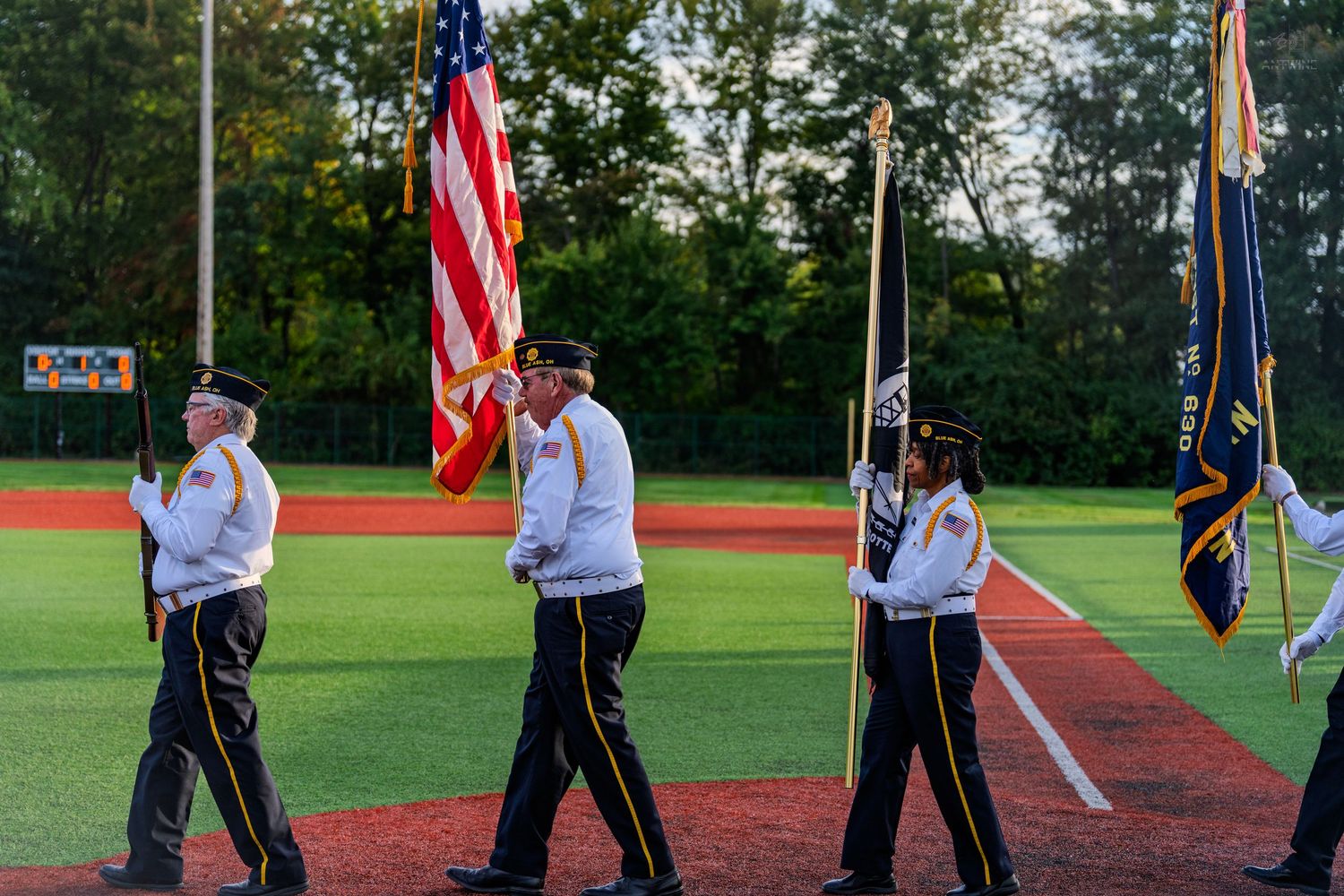 Veterans color guard marches with American and military flags on red clay baseball field at dusk.