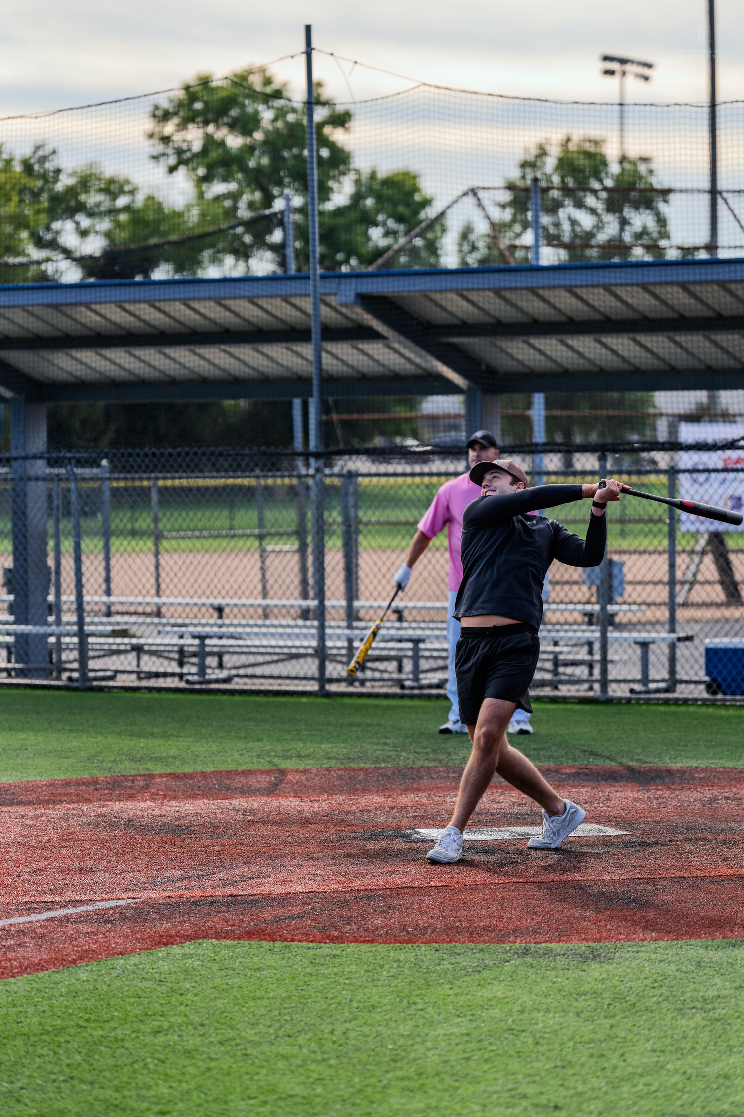 A baseball player in pink and black follows through on a swing at an outdoor ballfield with dugout and trees behind.
