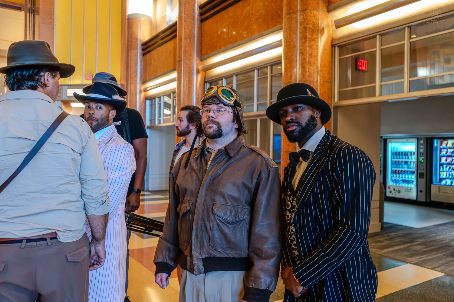 Group of men in steampunk costumes with hats and goggles posing in a building lobby near a vending machine.