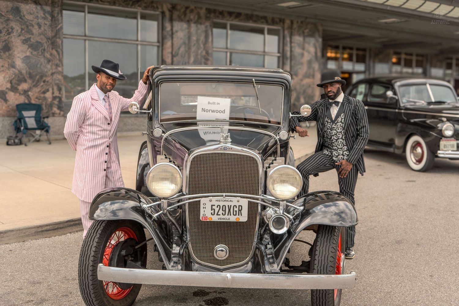 Two elegantly dressed people pose with a vintage 1930s classic car at an outdoor antique auto show.