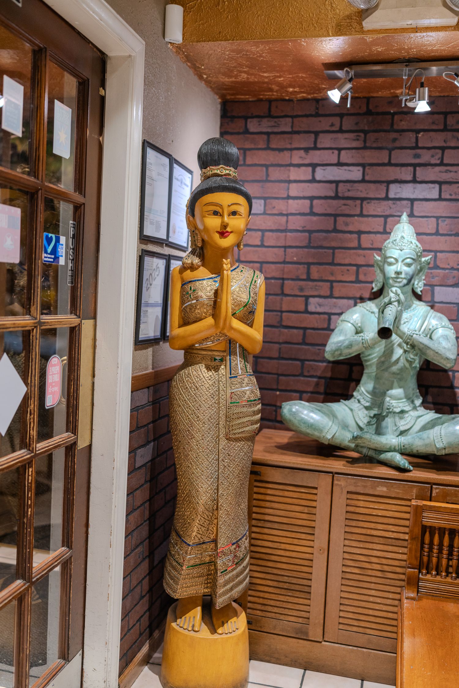 A golden Thai female statue in prayer pose stands beside a seated Buddha figurine in a brick-walled restaurant entrance.