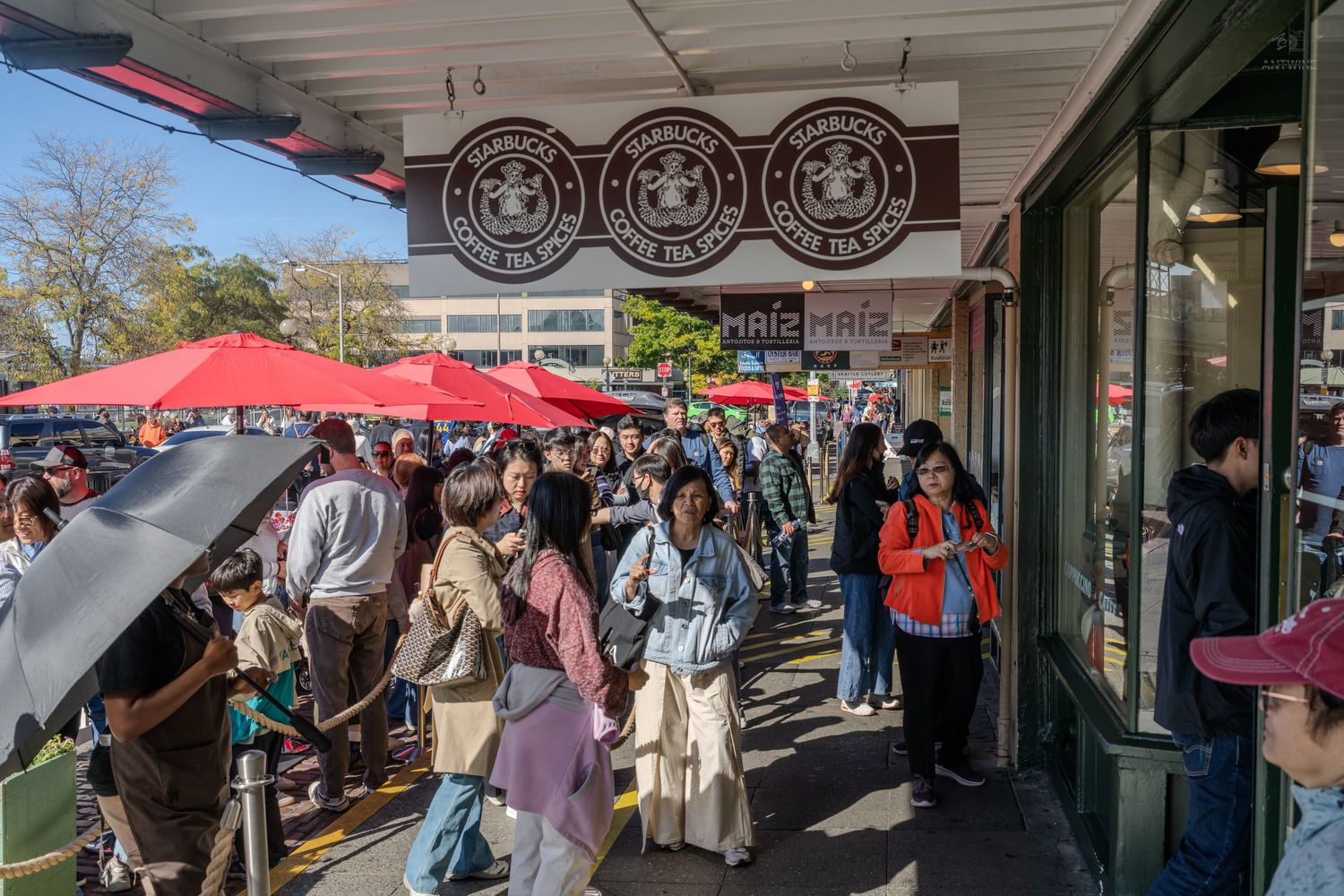 Large crowd queuing outside the original Starbucks store at Pike Place Market in Seattle with red umbrellas.