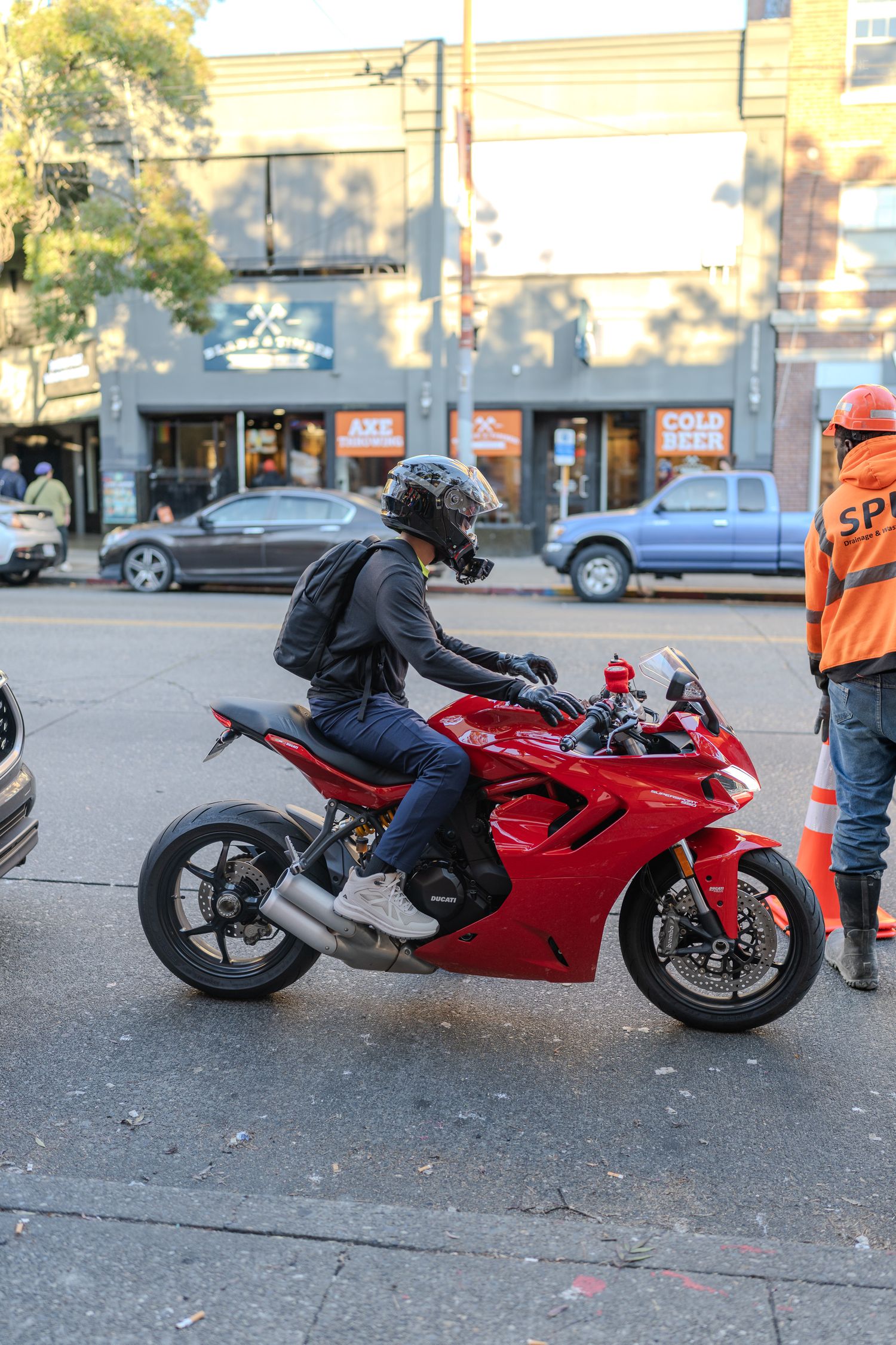 Red Ducati motorcycle ridden by helmeted rider on urban street with construction worker and shops in background.