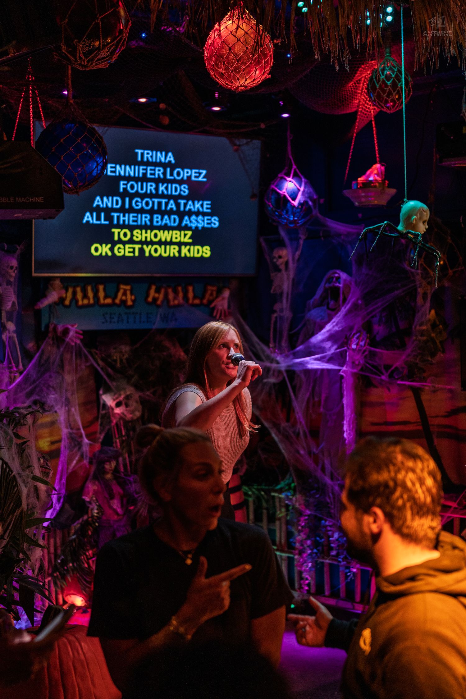 Woman singing karaoke at Hula Hula bar surrounded by Halloween decorations and colorful lights.