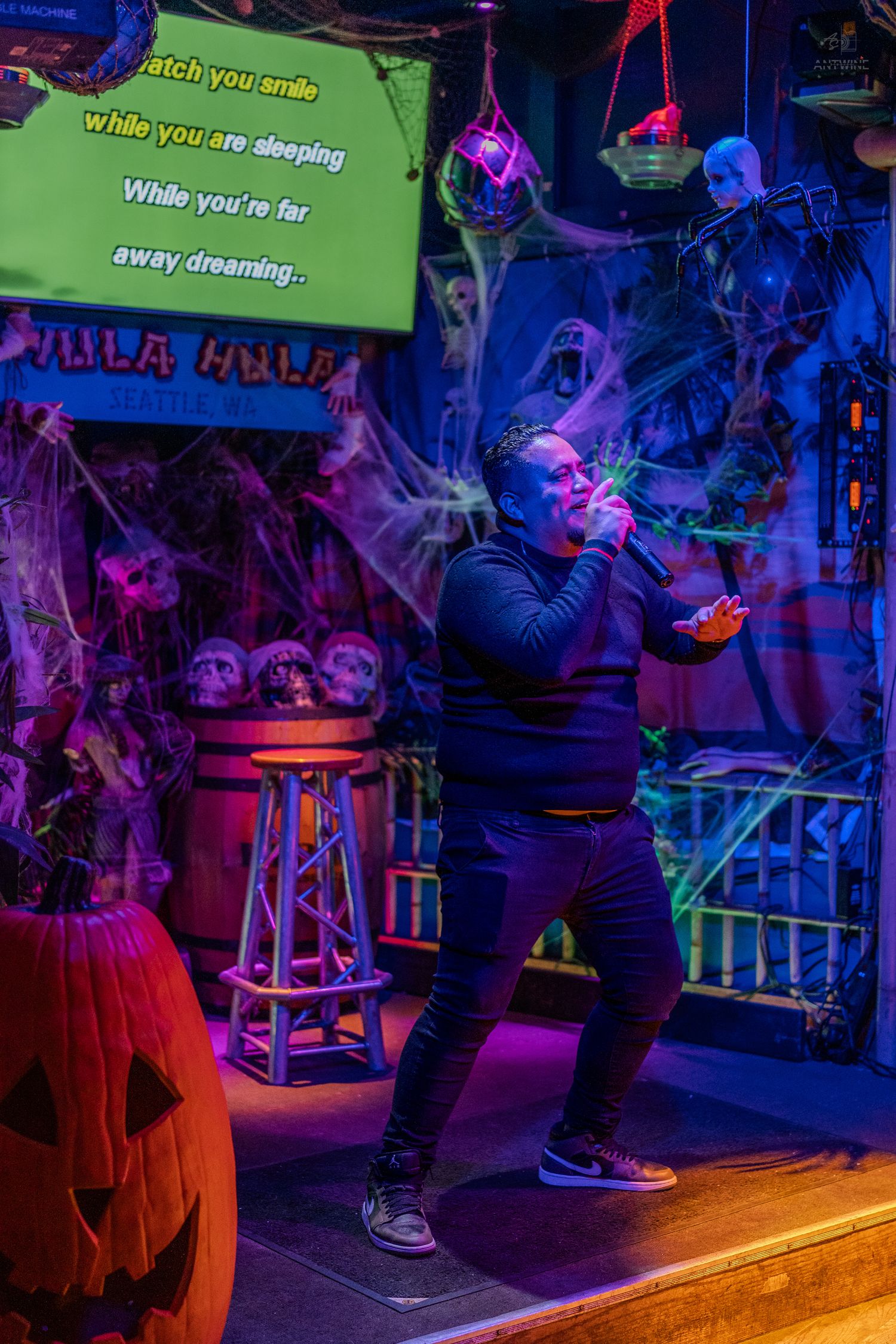 Man singing karaoke passionately on Halloween stage with cobwebs and skulls at Hula Hula bar.