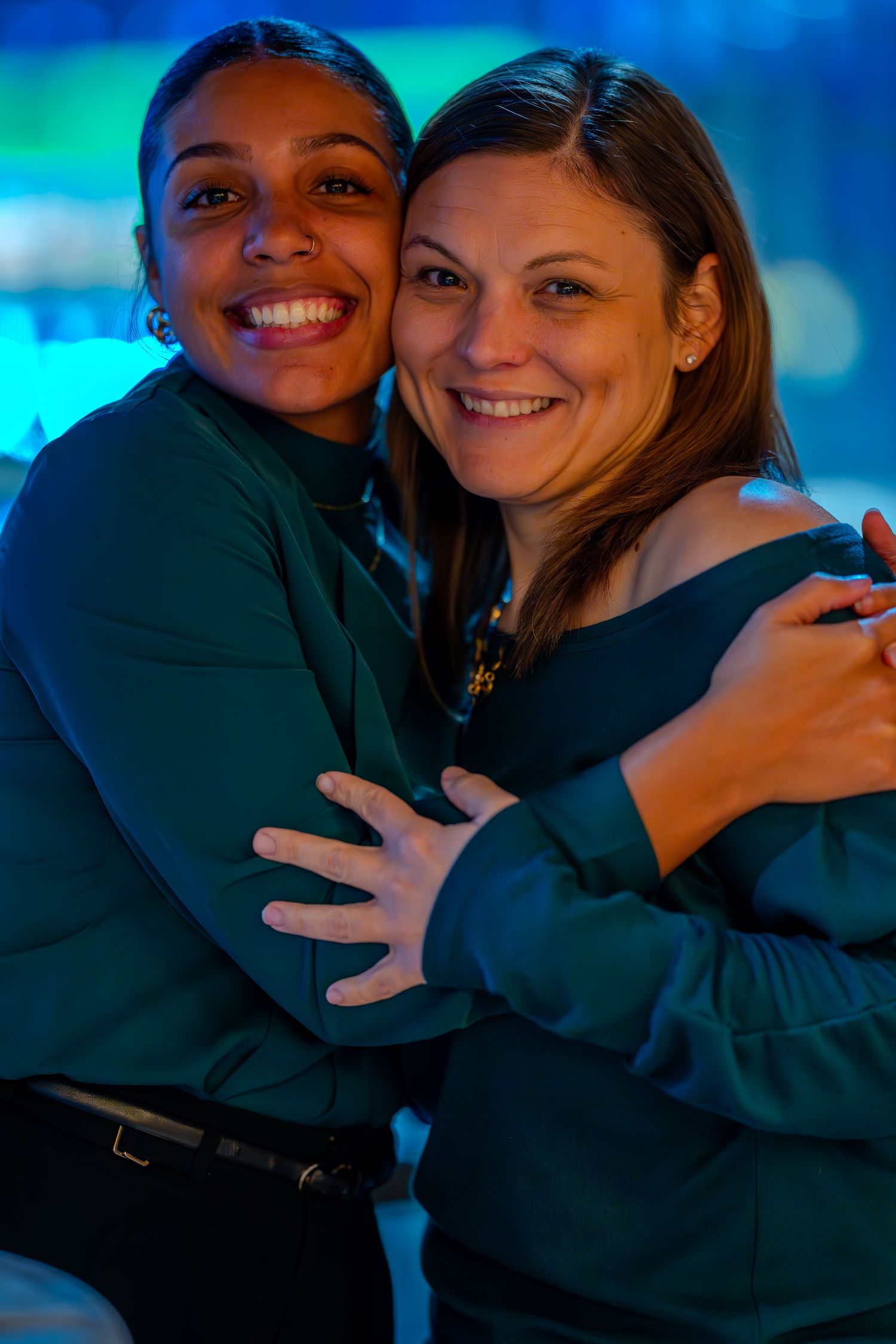 Two women smiling and hugging warmly, wearing matching green tops in a blue-lit setting.
