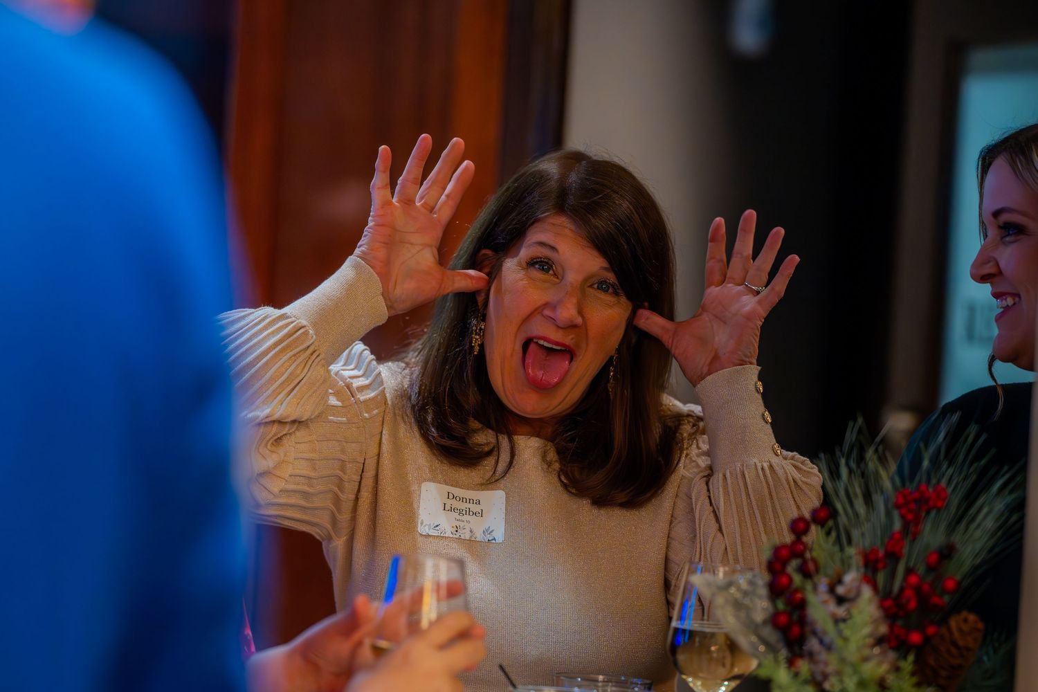 Woman with hands raised in excitement at a social gathering event, wearing a name tag and beige sweater.