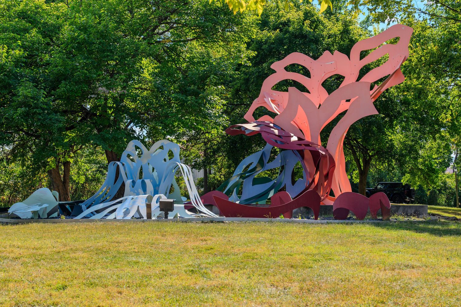 Colorful abstract metal sculptures displayed on green grass surrounded by lush trees in an outdoor park setting.
