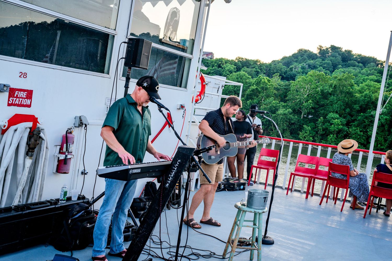 Two musicians playing keyboard and guitar on a riverboat deck surrounded by lush green trees at sunset.
