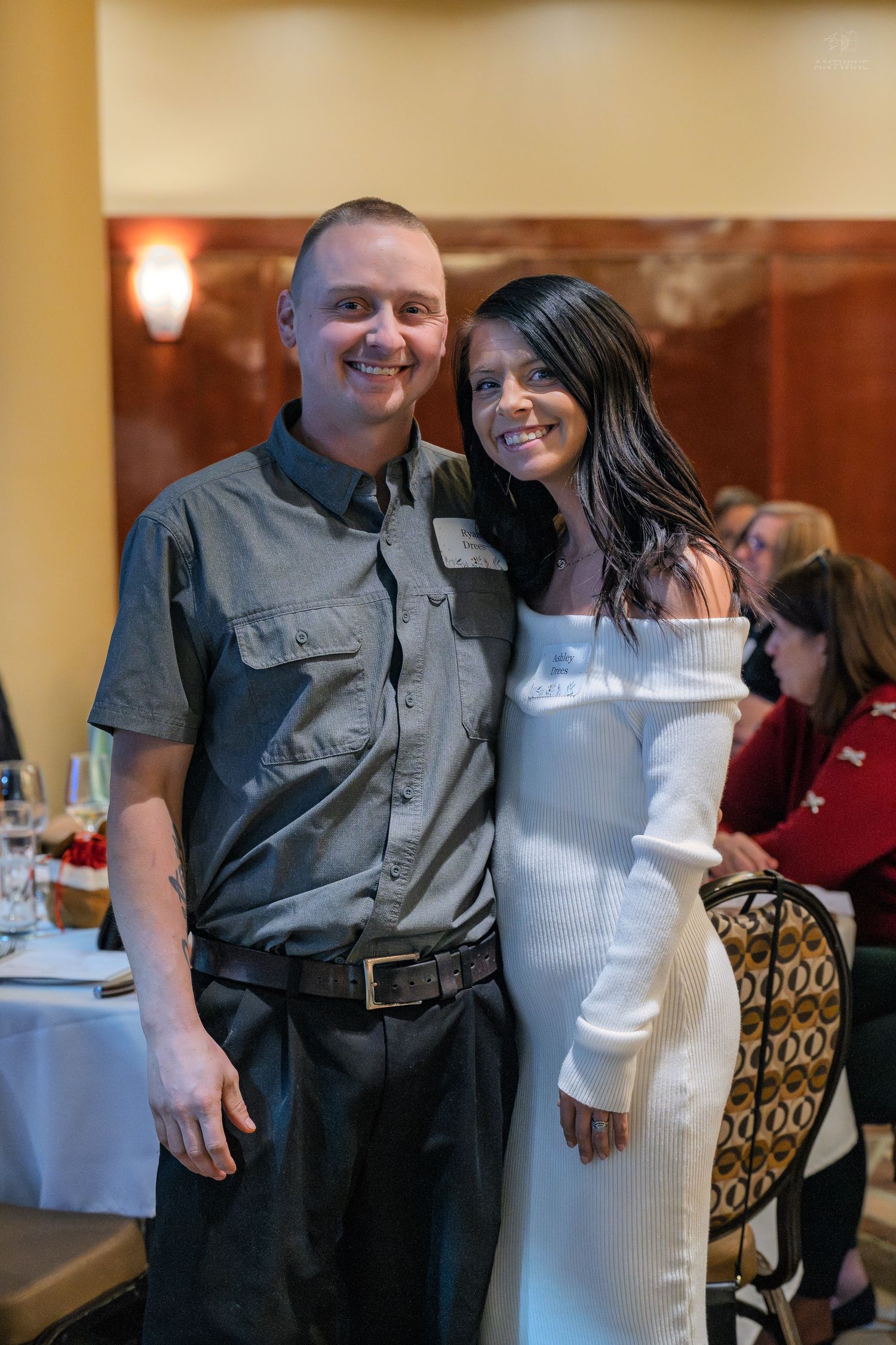 A smiling couple poses together at a restaurant, man in gray shirt and woman in white off-shoulder dress.