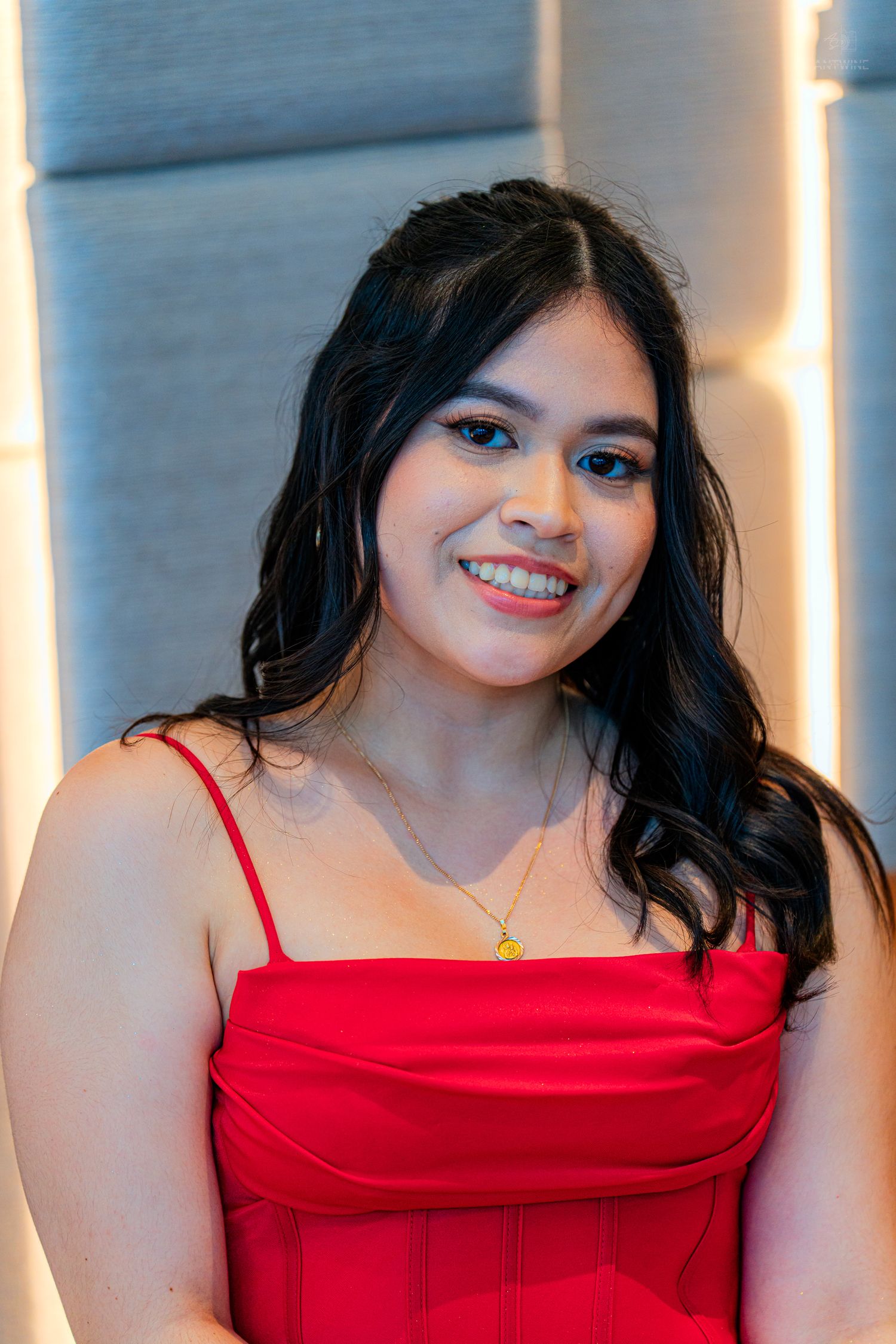 Smiling young woman in red dress with gold necklace posing indoors.