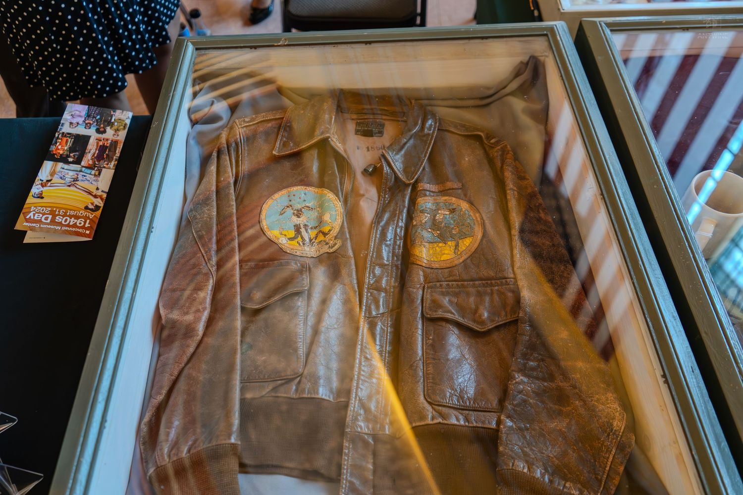 Vintage WWII leather bomber jacket with painted patches displayed in a glass museum exhibit case.