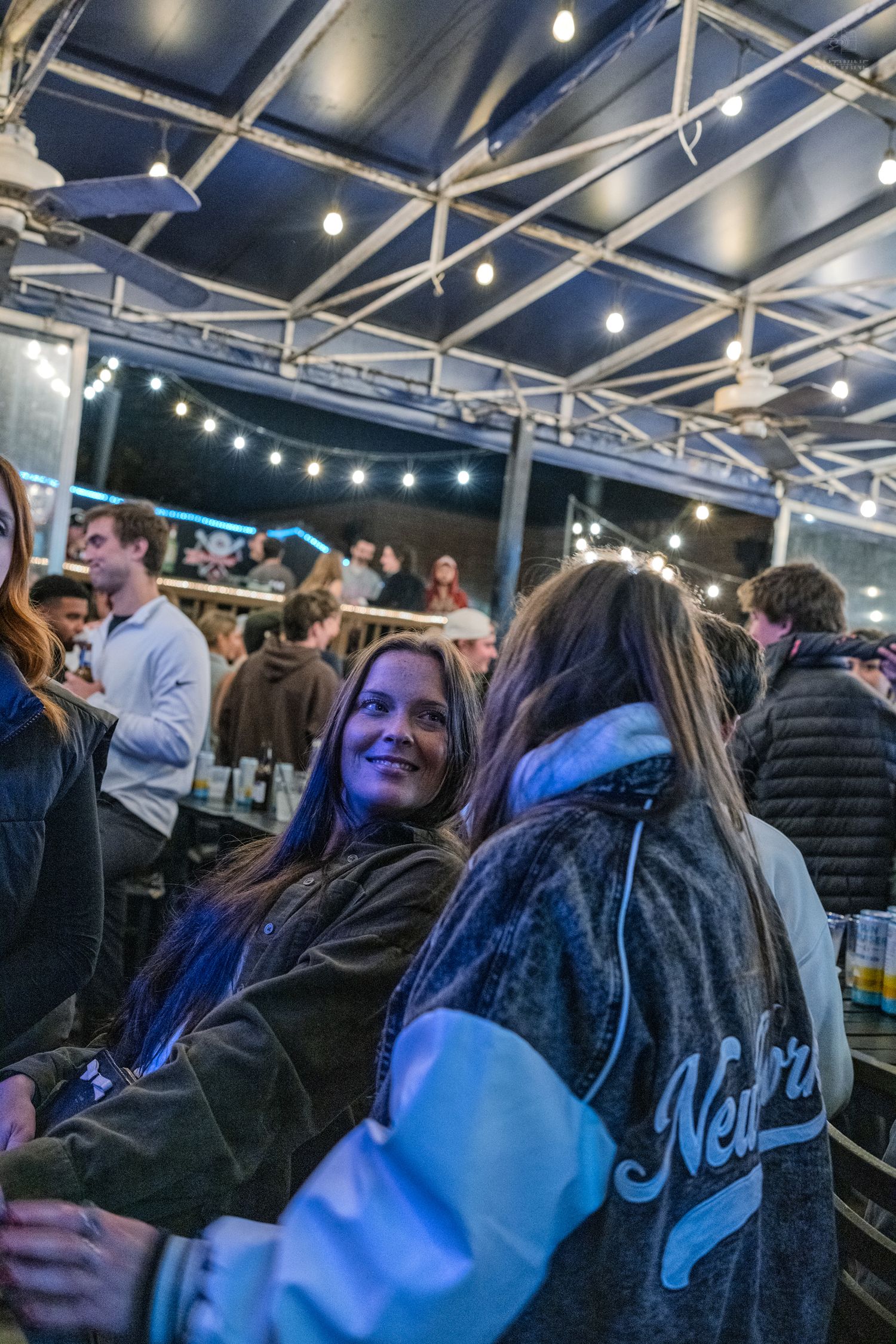Young woman with long dark hair smiling at a lively outdoor bar event under string lights at night.