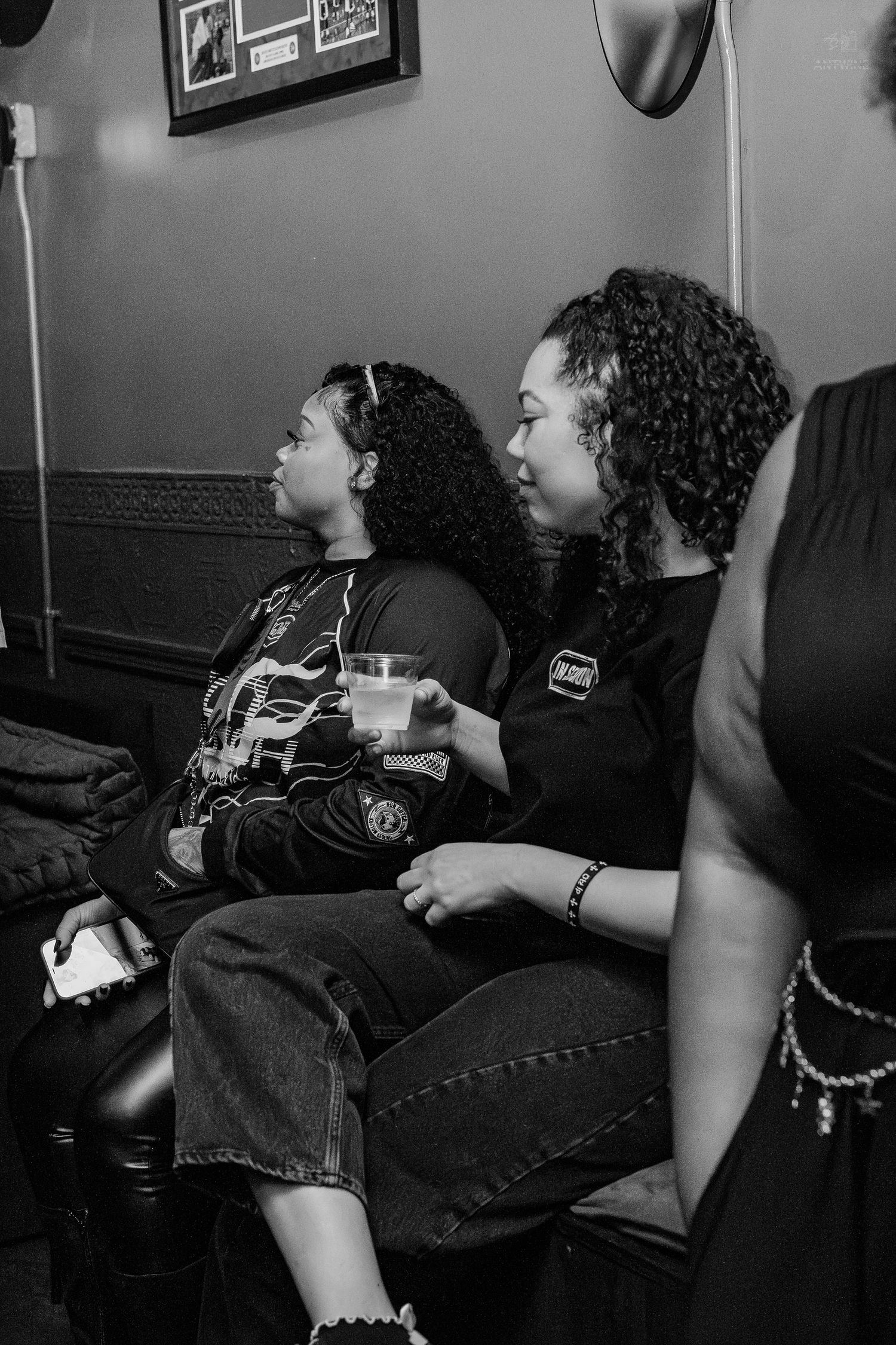Black and white photo of two young women with curly hair sitting together, one holding a drink, in a dimly lit room.