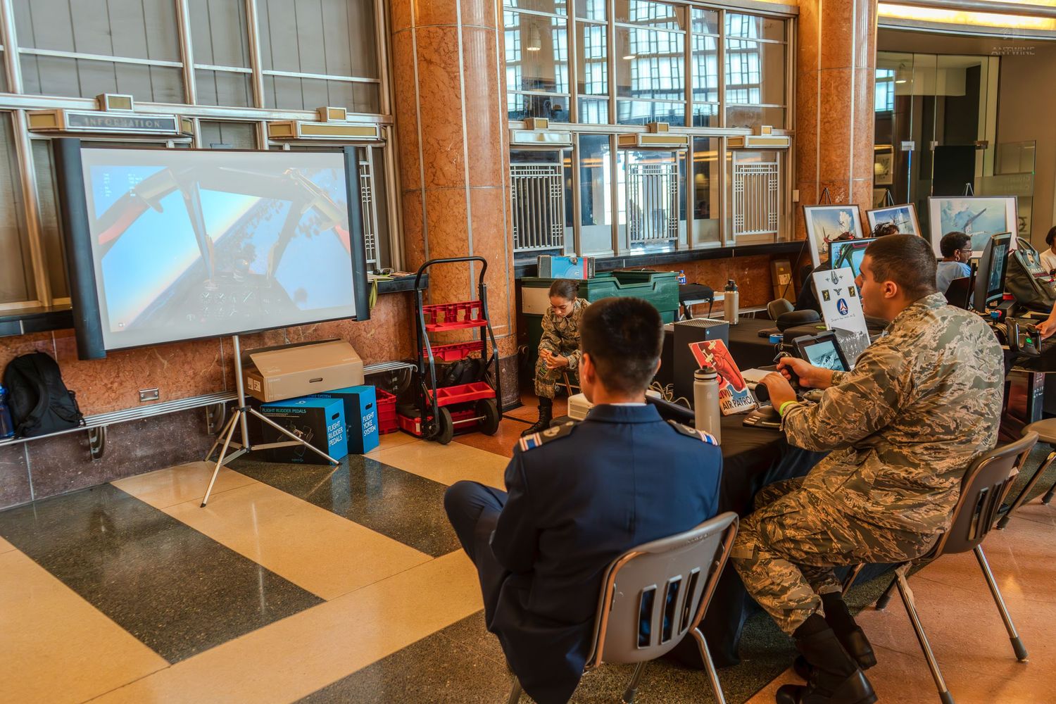 Military personnel and visitors watch a presentation on a projector screen inside a building lobby.
