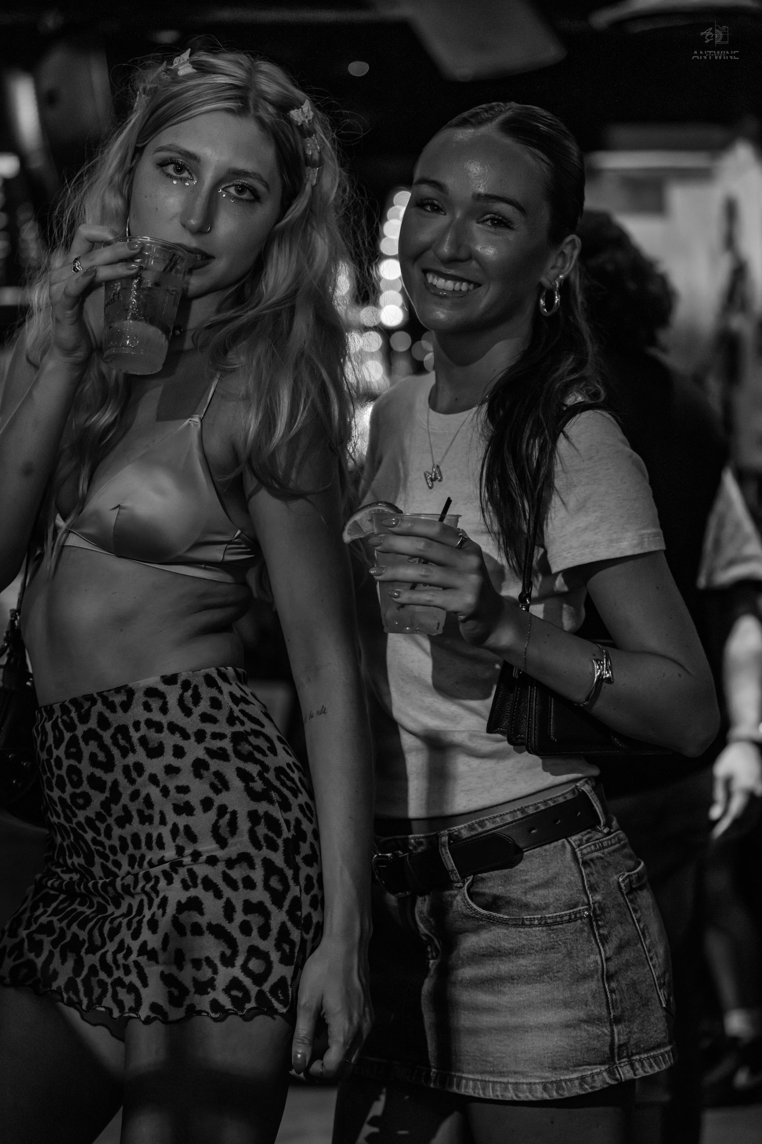 Two women smiling and holding drinks at a party, one in a leopard print skirt, captured in black and white.