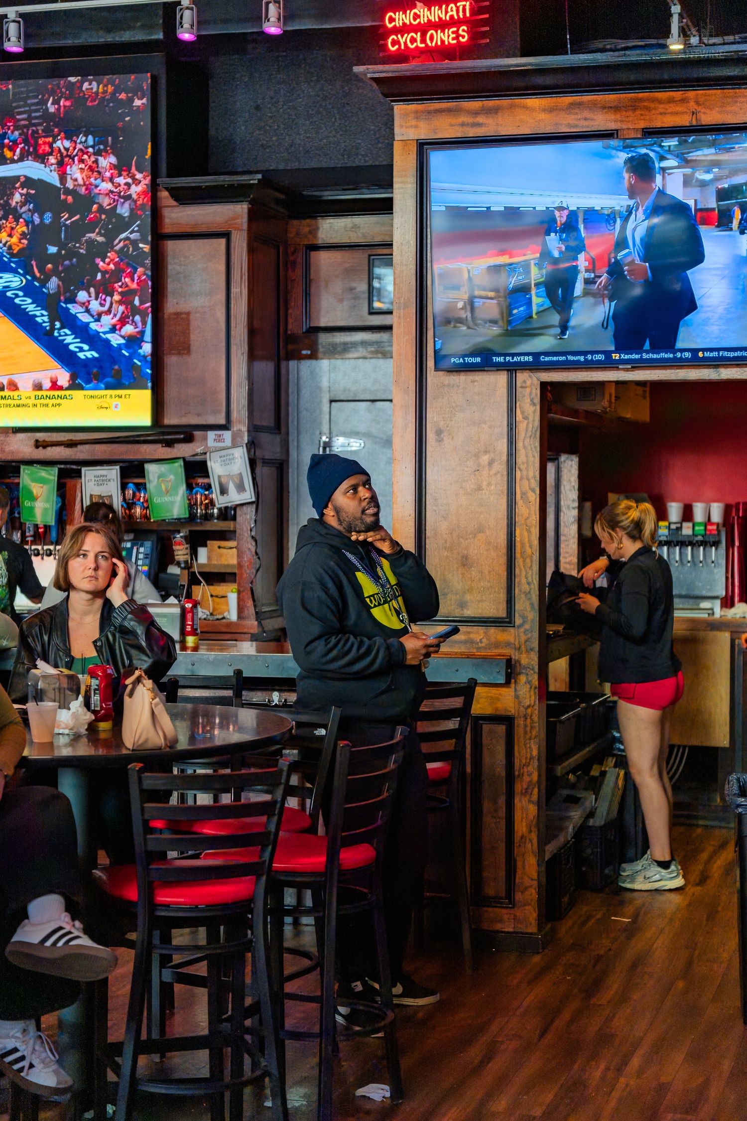 Patrons relaxing inside a sports bar with TVs, high-top tables, and a Cincinnati Cyclones neon sign.
