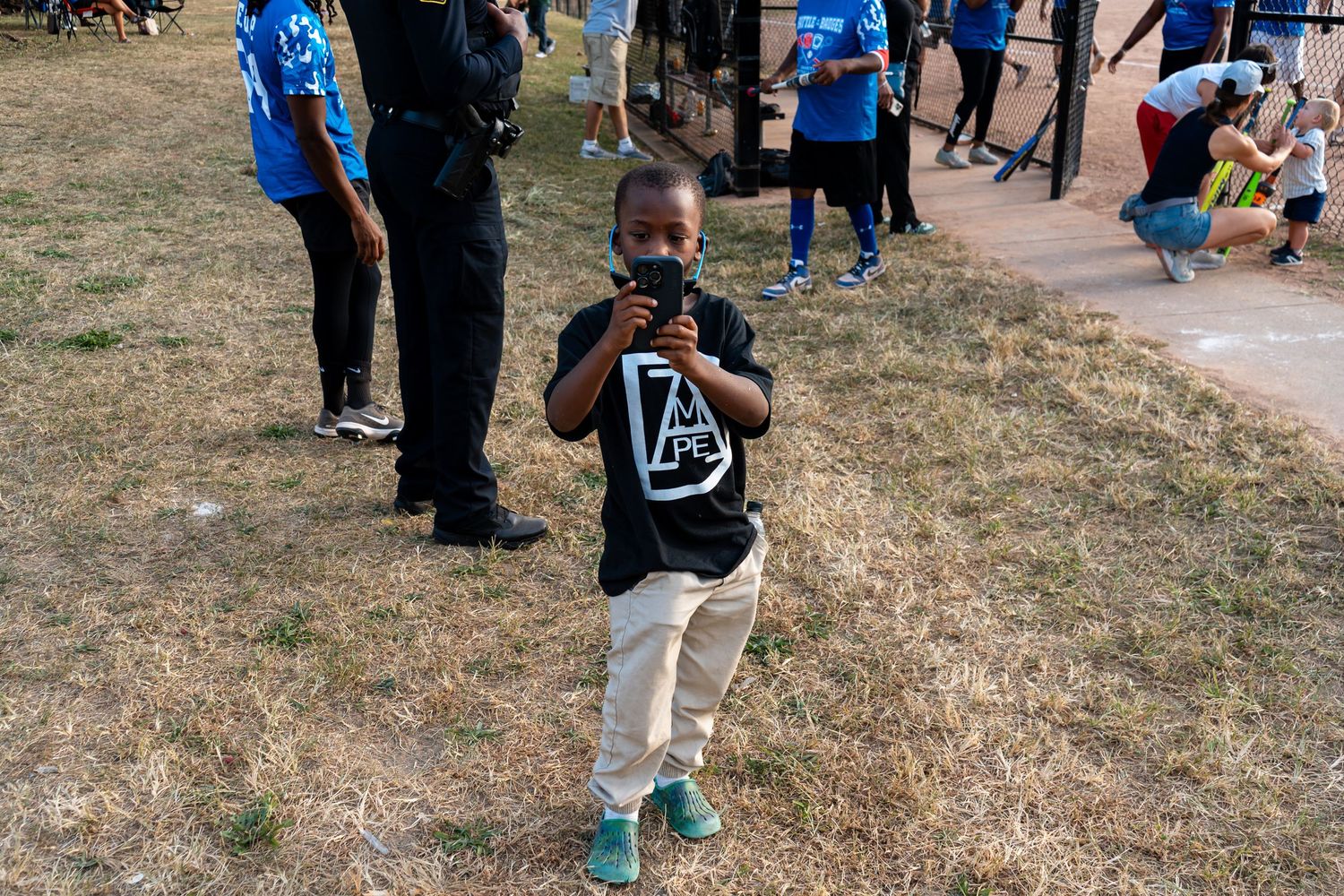 Young Black boy in a black t-shirt walking on dry grass at an outdoor community event.