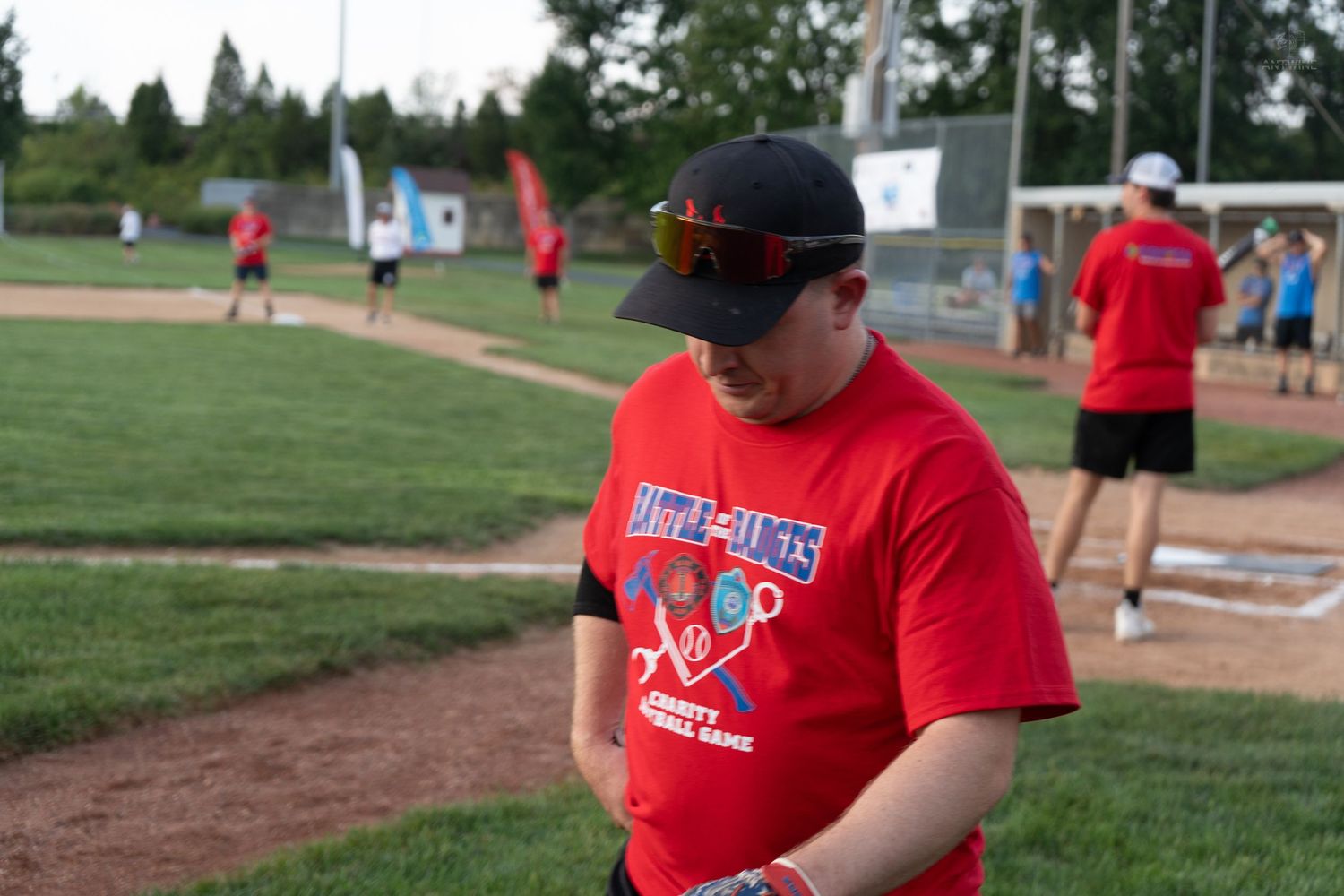 A man in a red charity softball league t-shirt and black cap stands on the baseball field during a game.