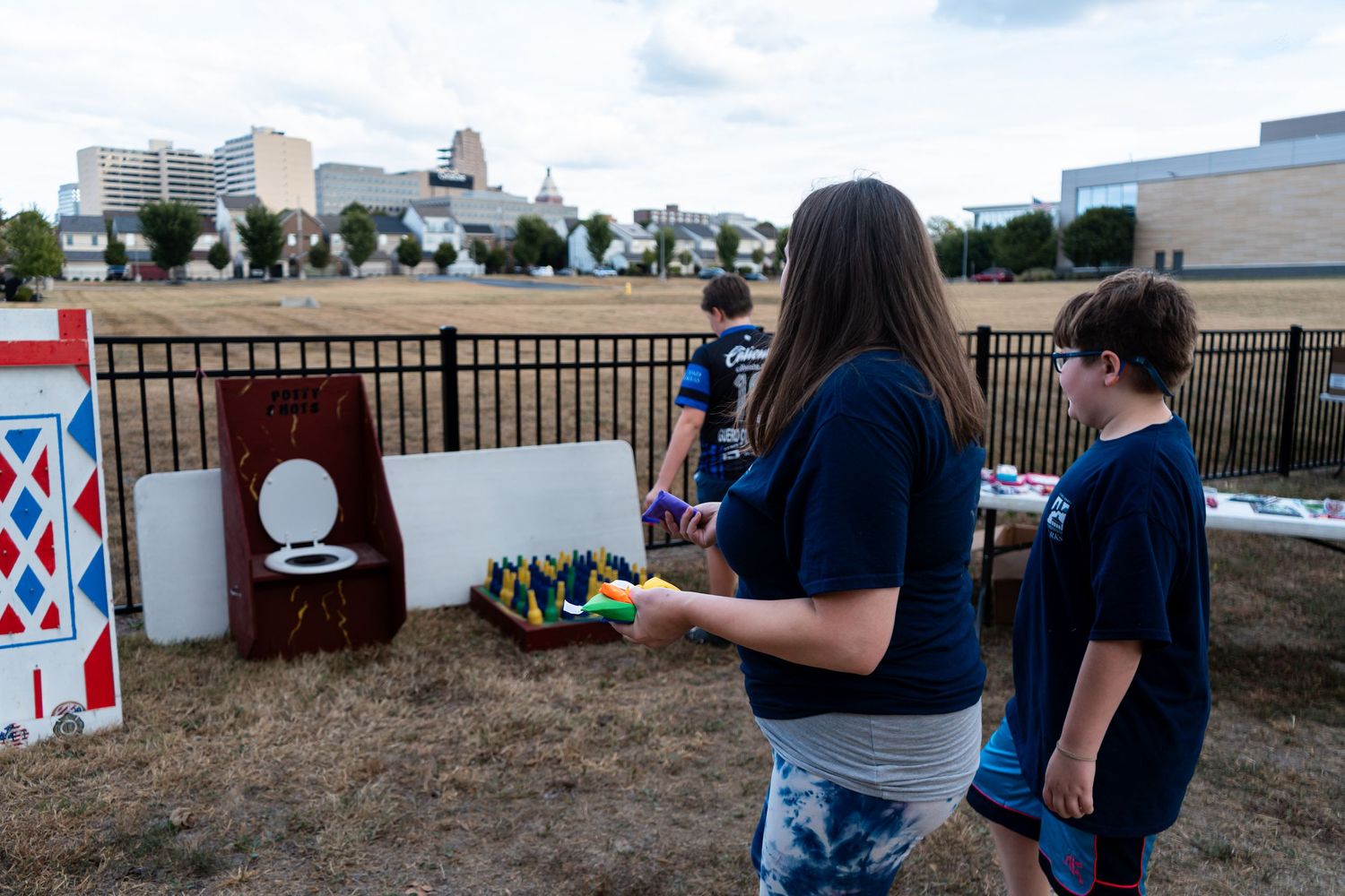 Children play colorful lawn games at an outdoor softball charity event near a fenced field.