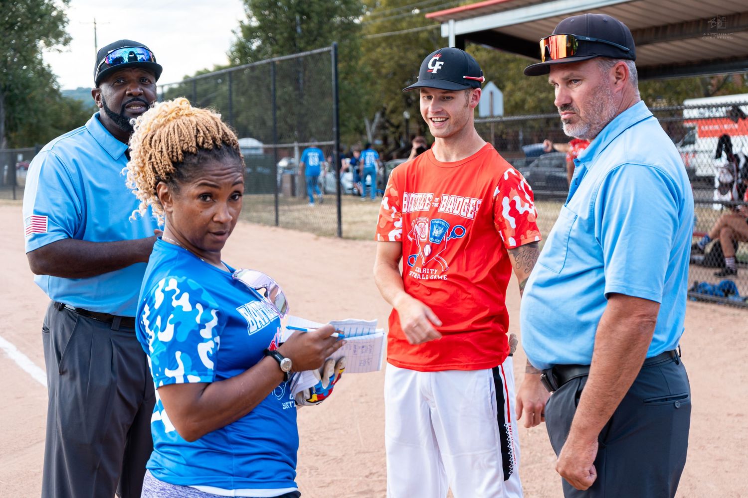 Umpires and players in blue and red uniforms discuss game details on a softball field during an outdoor event.