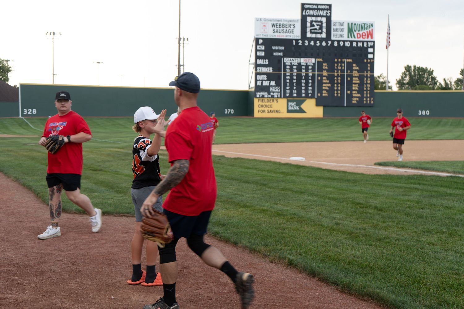 Adults and children in red shirts play baseball on a professional field with green scoreboard.