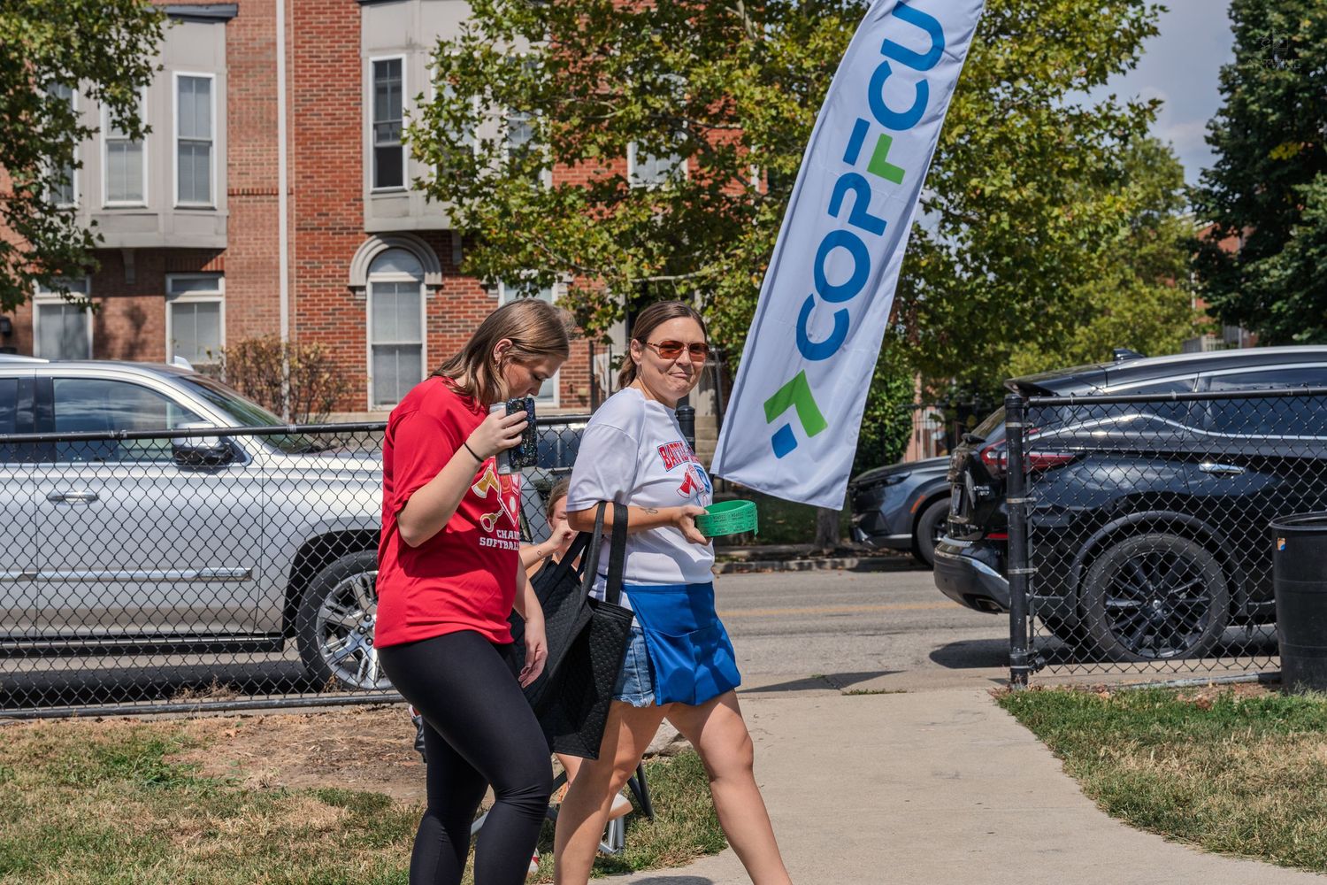 Two women walking on a sidewalk near a COPCU banner during an outdoor community sporting event on a sunny day.