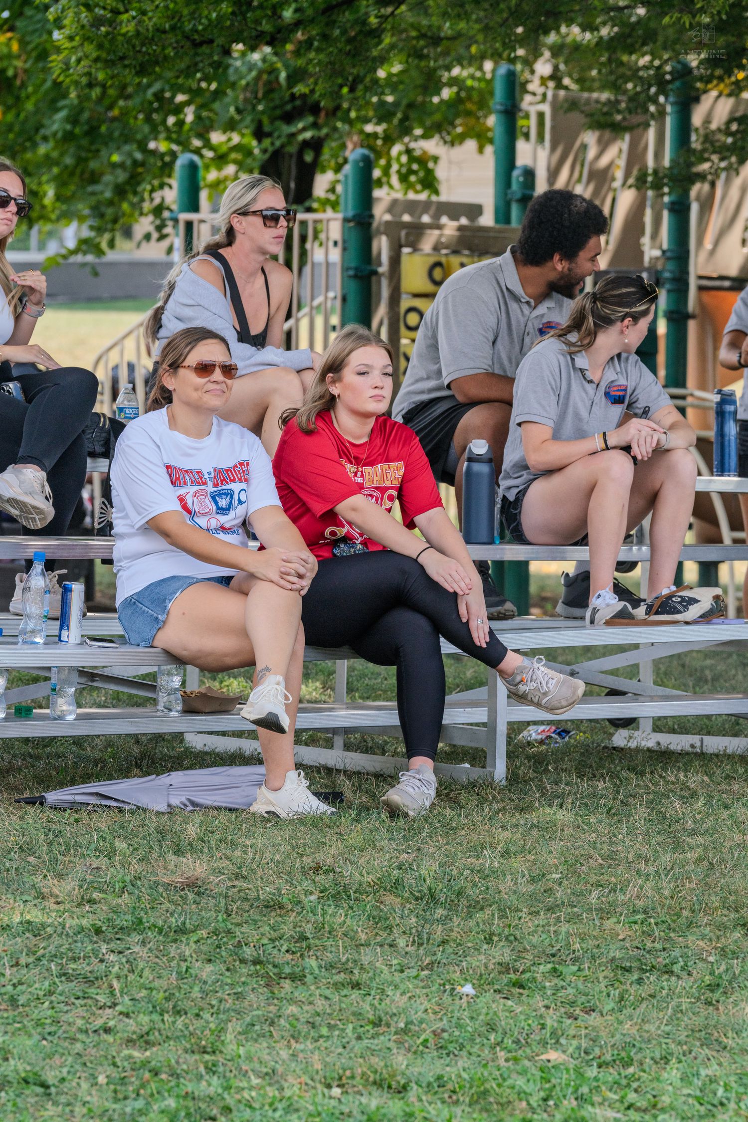 Spectators sitting on bleachers watching a Battle of the Badges charity softball game outdoors.