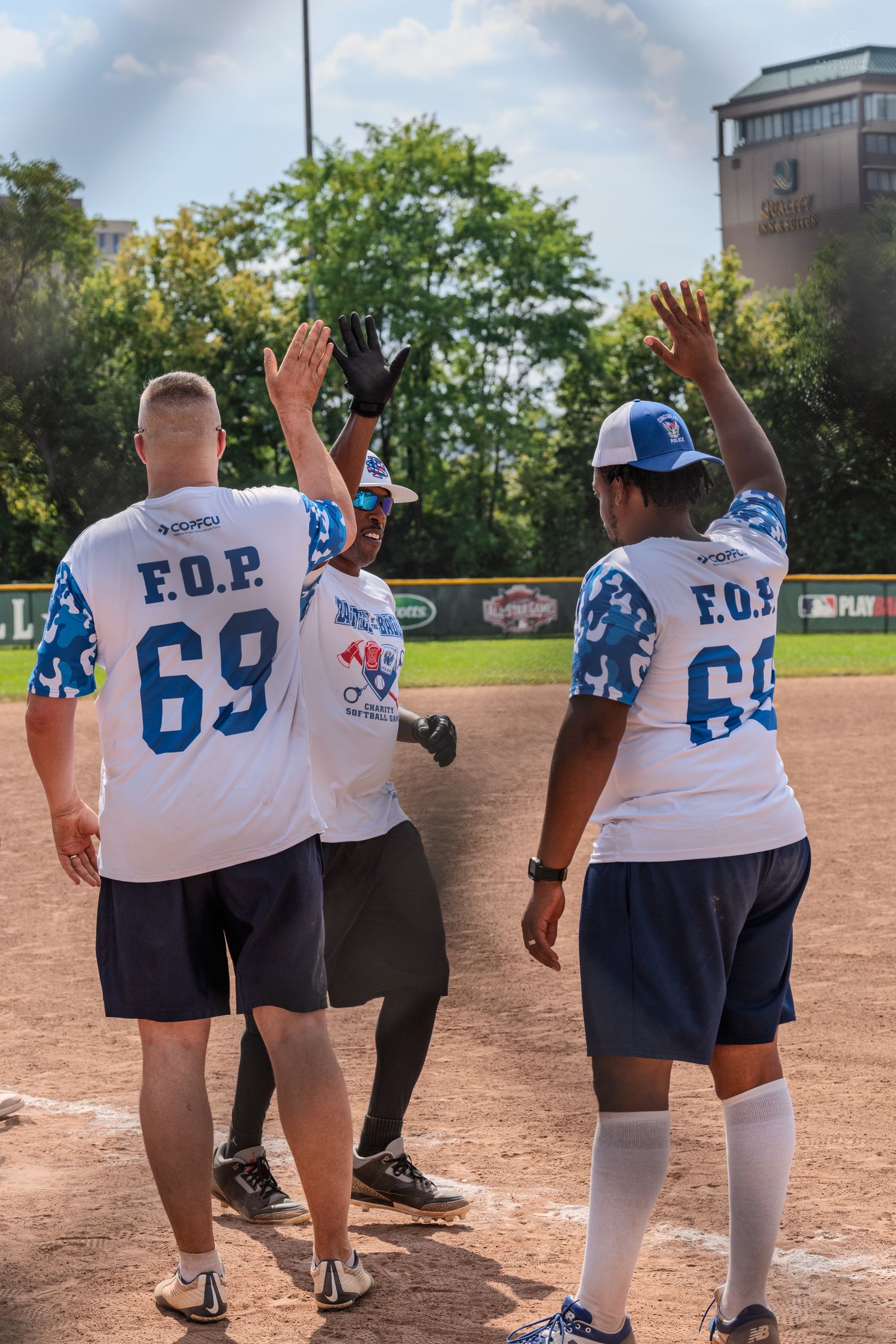 Three F.O.P. softball players celebrate with high-fives on the baseball diamond during a sunny game.