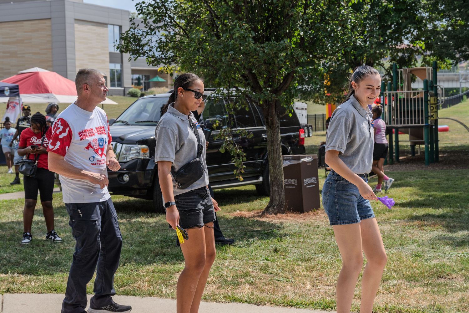 Three people walking on a sunny sidewalk at an outdoor community event near a playground.