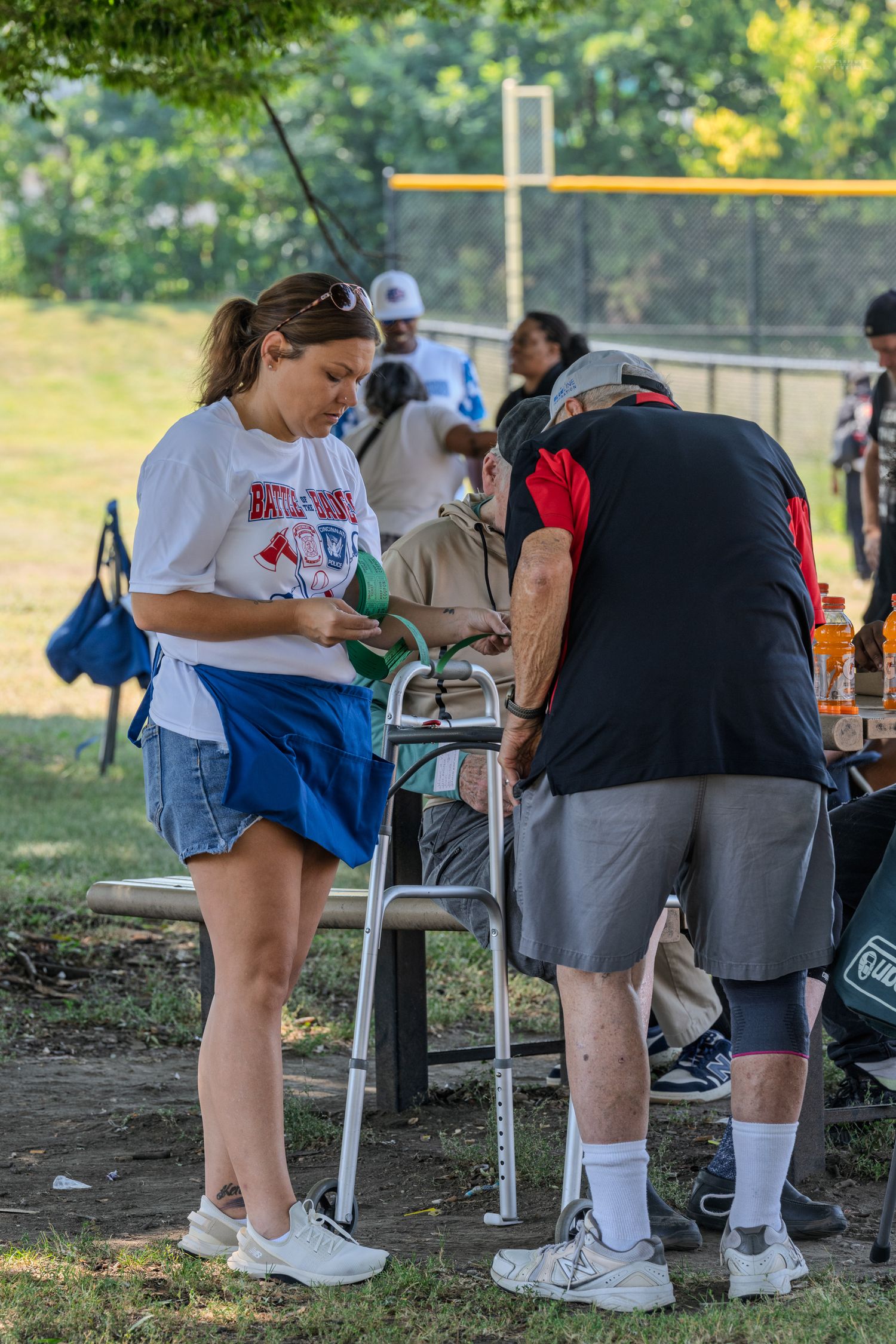 Woman in white shirt assists elderly man with walker at outdoor baseball field picnic area.