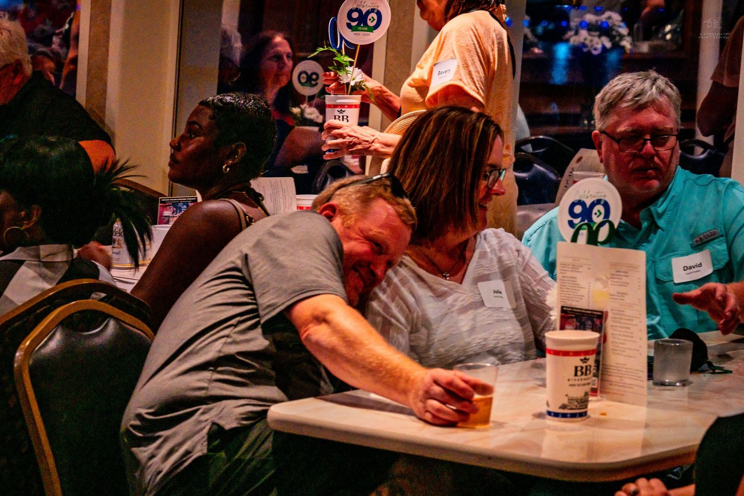 People socializing at an indoor party event, with a man resting his head on a woman's shoulder at a table.