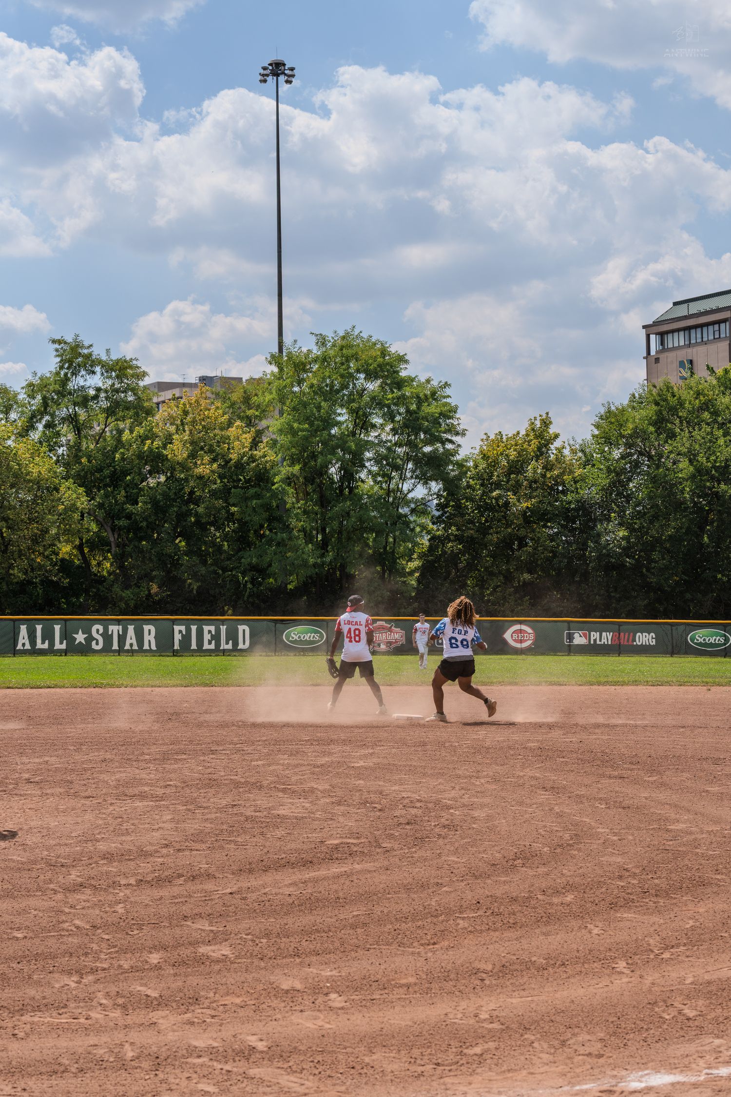 Two players run the bases on an All-Star Field during a sunny softball game.
