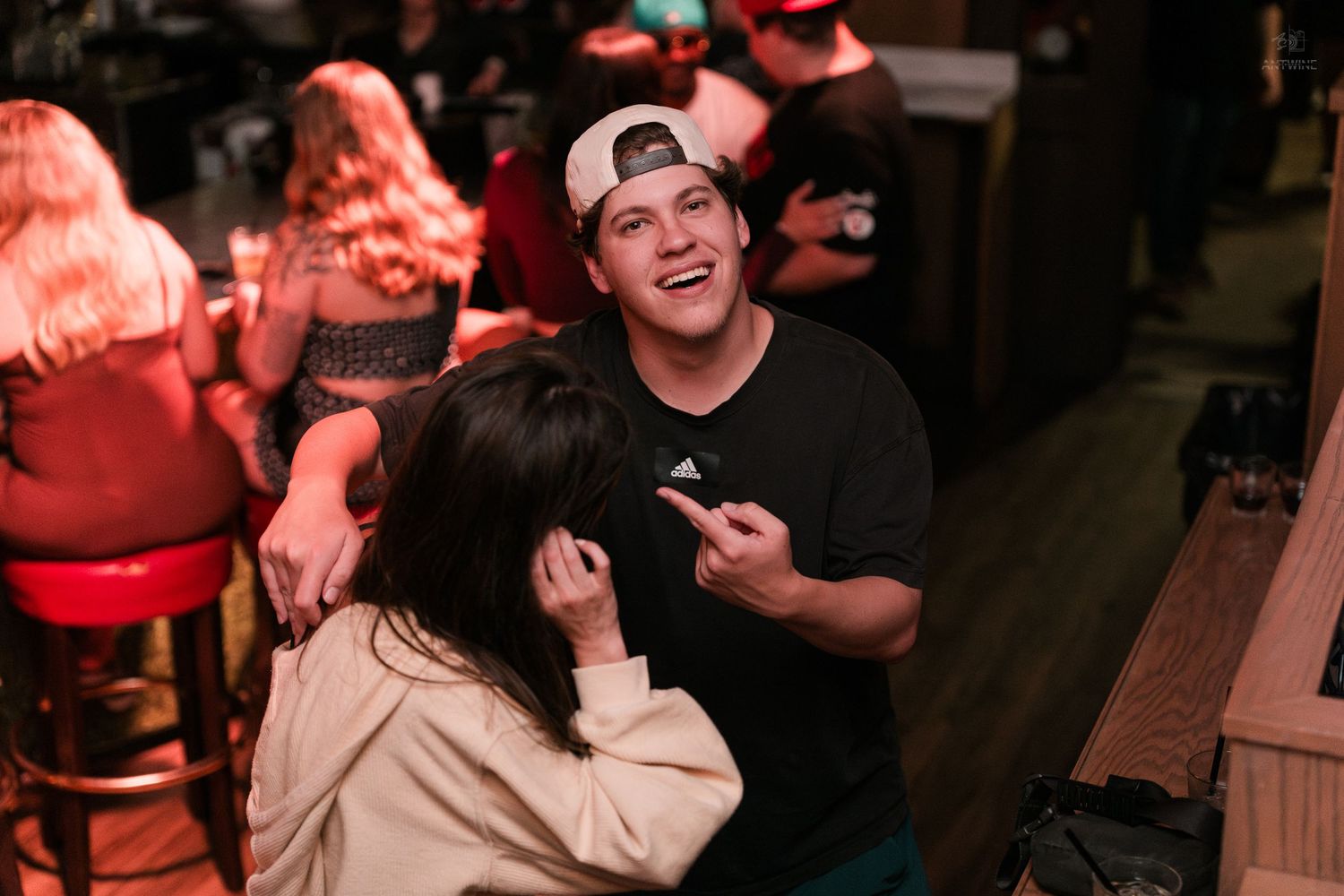 A cheerful young man in a backwards Adidas cap smiles and gestures while talking to a woman at a bar.
