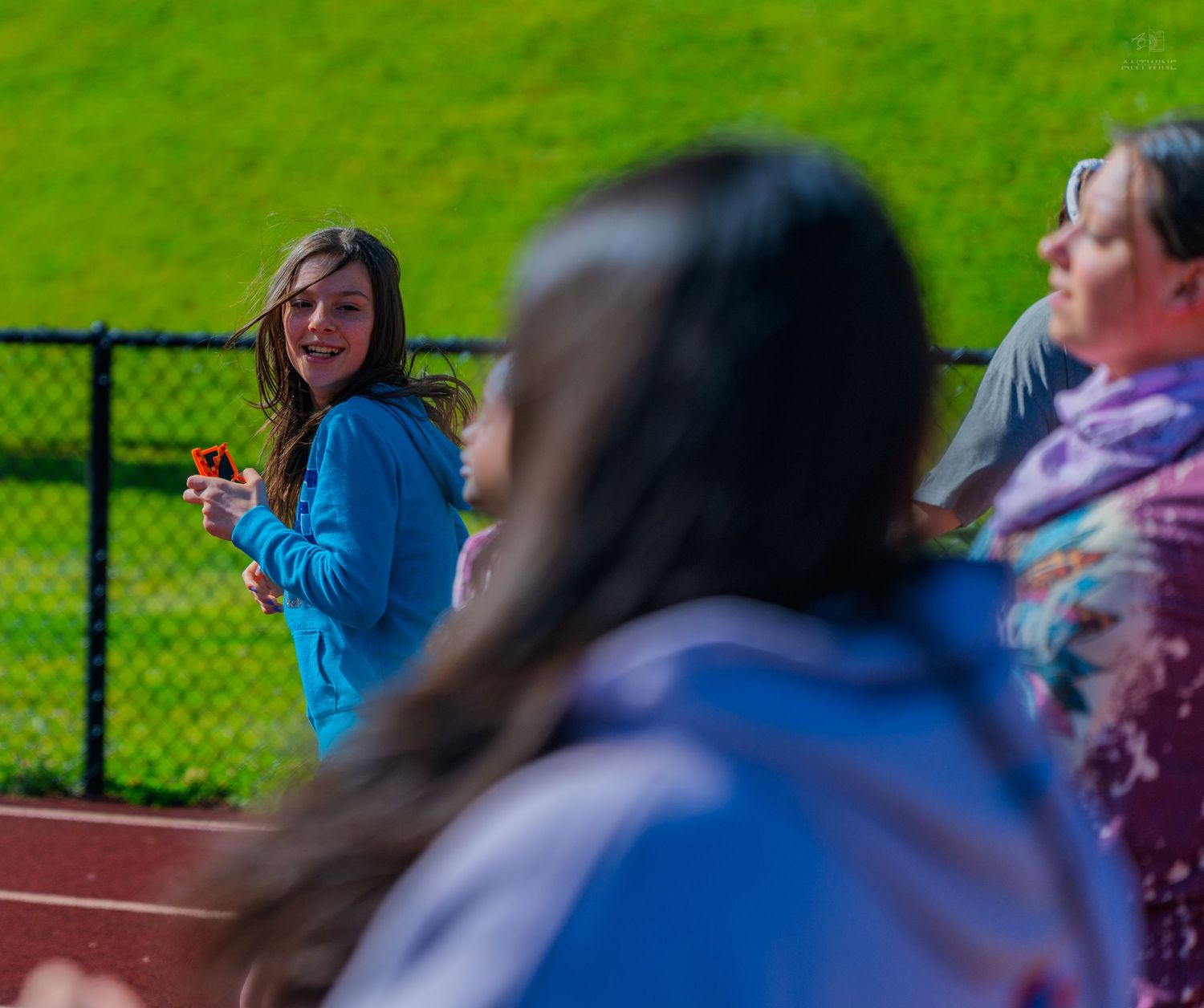 A smiling girl in a blue hoodie holds a small item outdoors near a track field with green grass in the background.