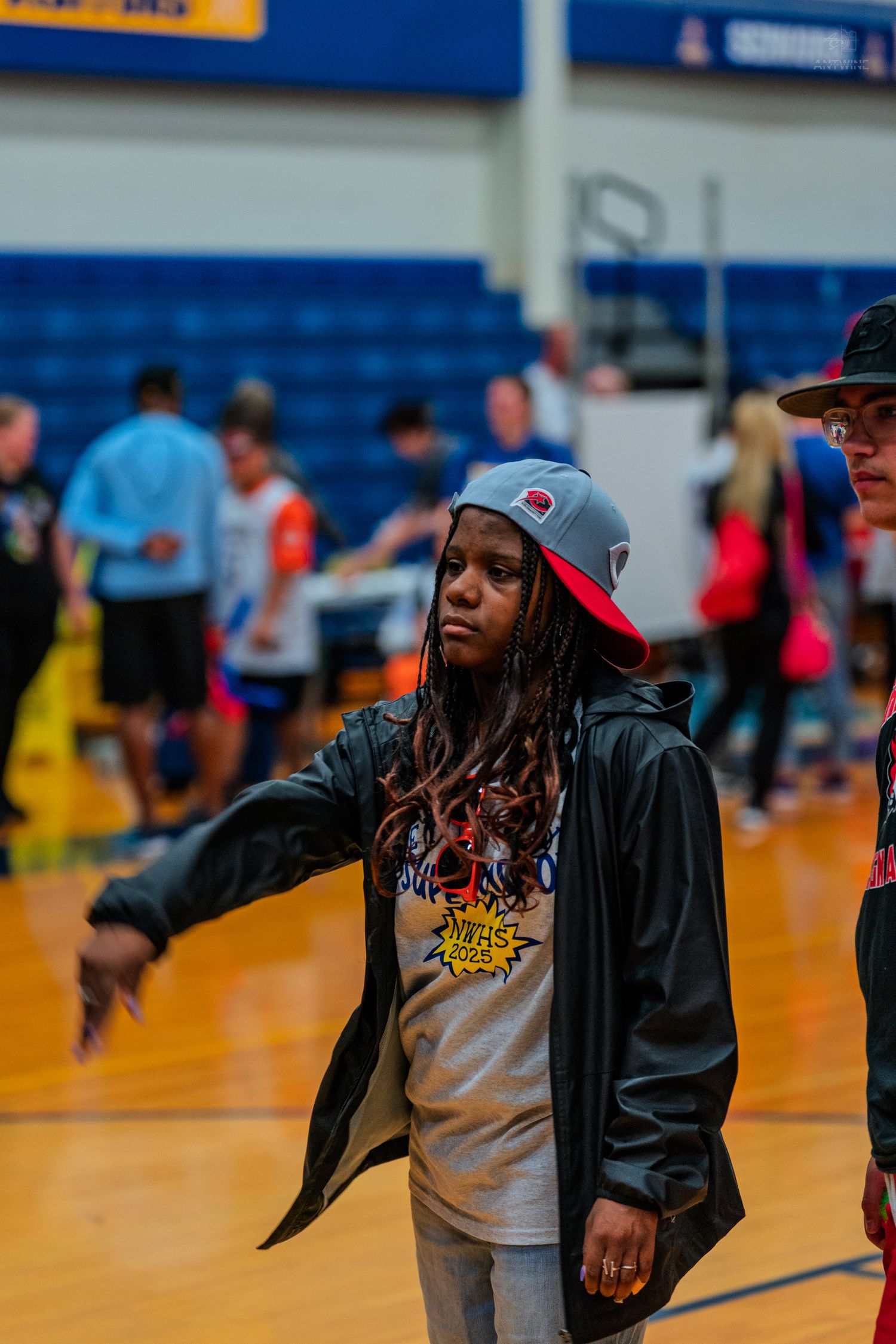 Young woman with dreadlocks wearing a snapback cap and black jacket gestures on a basketball court.