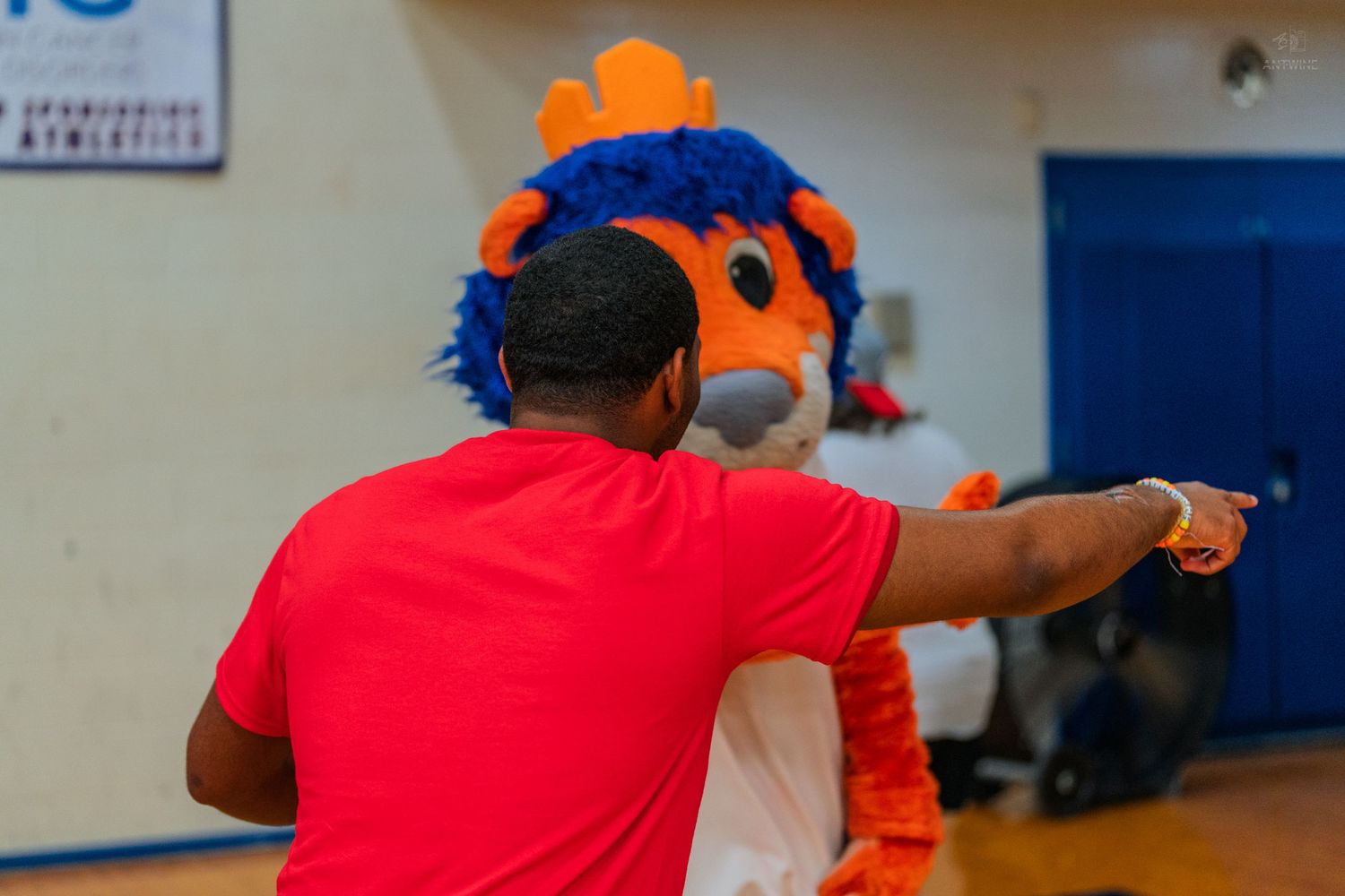 A man in a red shirt points directions alongside a colorful orange and blue lion mascot in a gymnasium.
