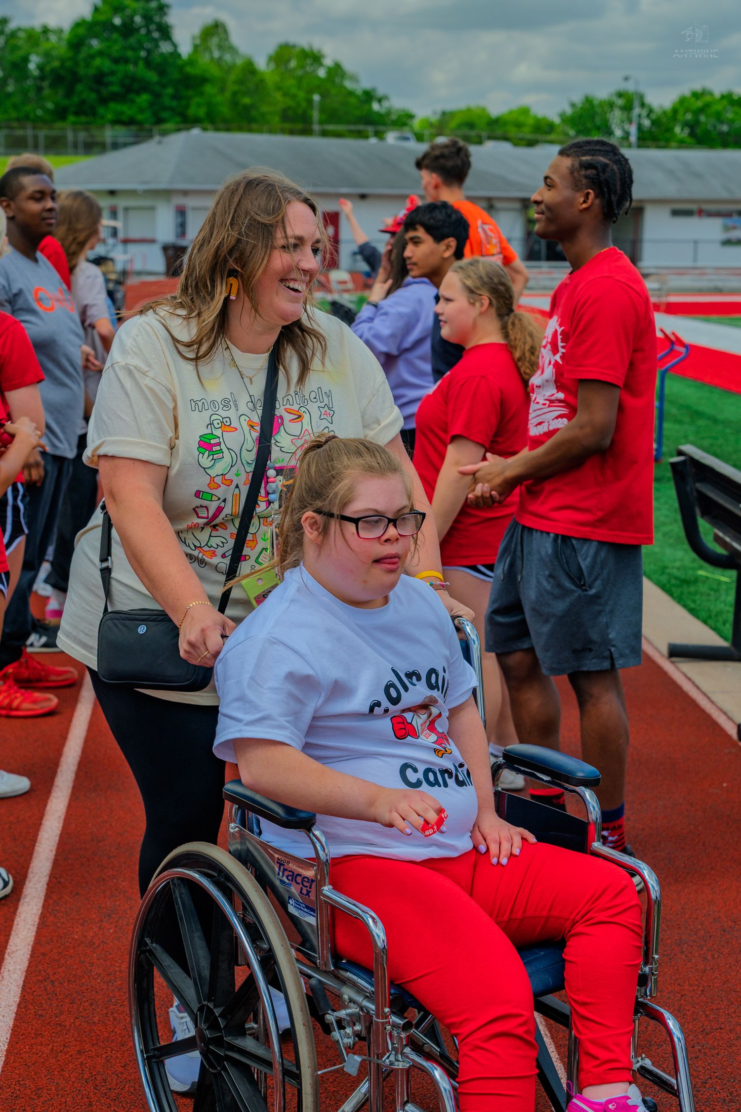 A smiling volunteer pushes a young athlete in a wheelchair on a red track at a Special Olympics event.