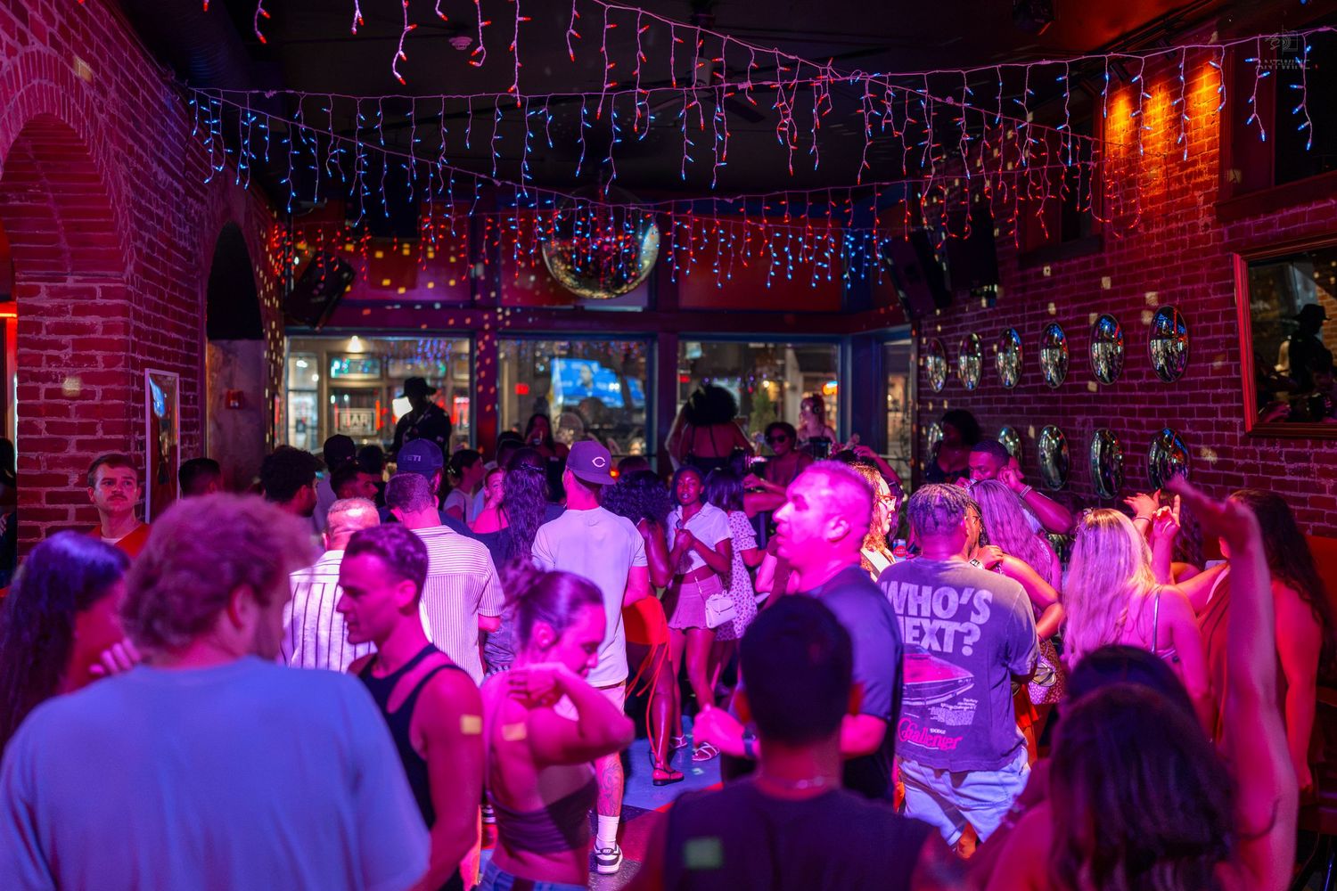 Crowded nightclub dance floor with colorful pink and purple lights, string lights on brick walls, and energetic partygoers.