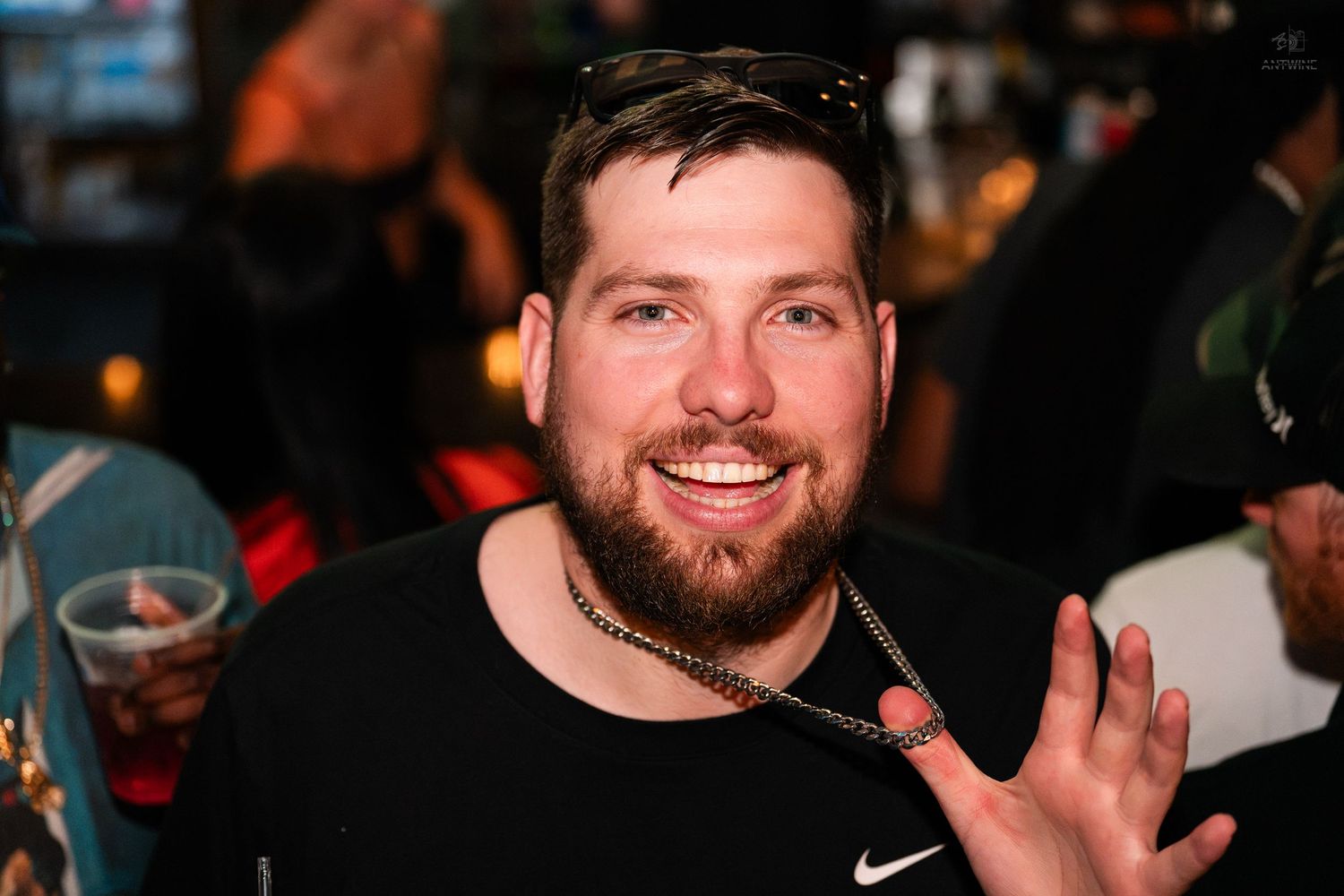 Smiling bearded man in black Nike shirt at lively bar or nightclub event.