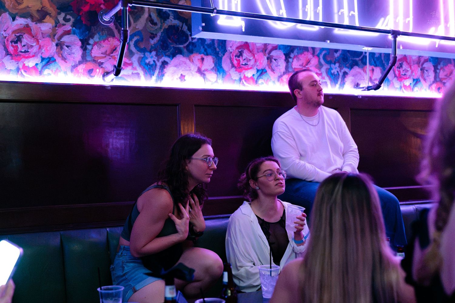 Group of friends relaxing in a nightclub booth under purple neon lights and floral decor.
