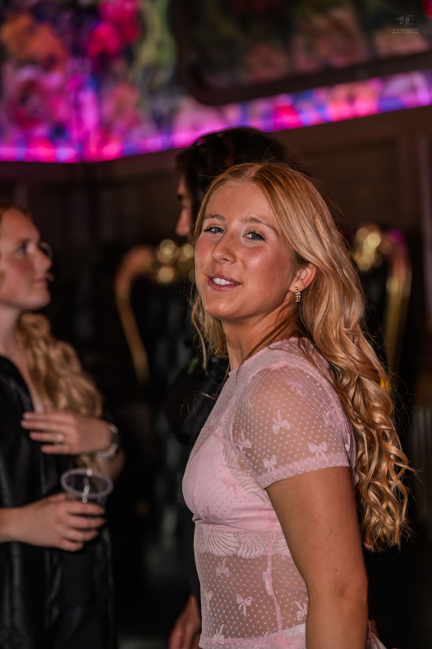 Smiling blonde woman in pink lace dress enjoys a nightclub event under colorful ambient lighting.