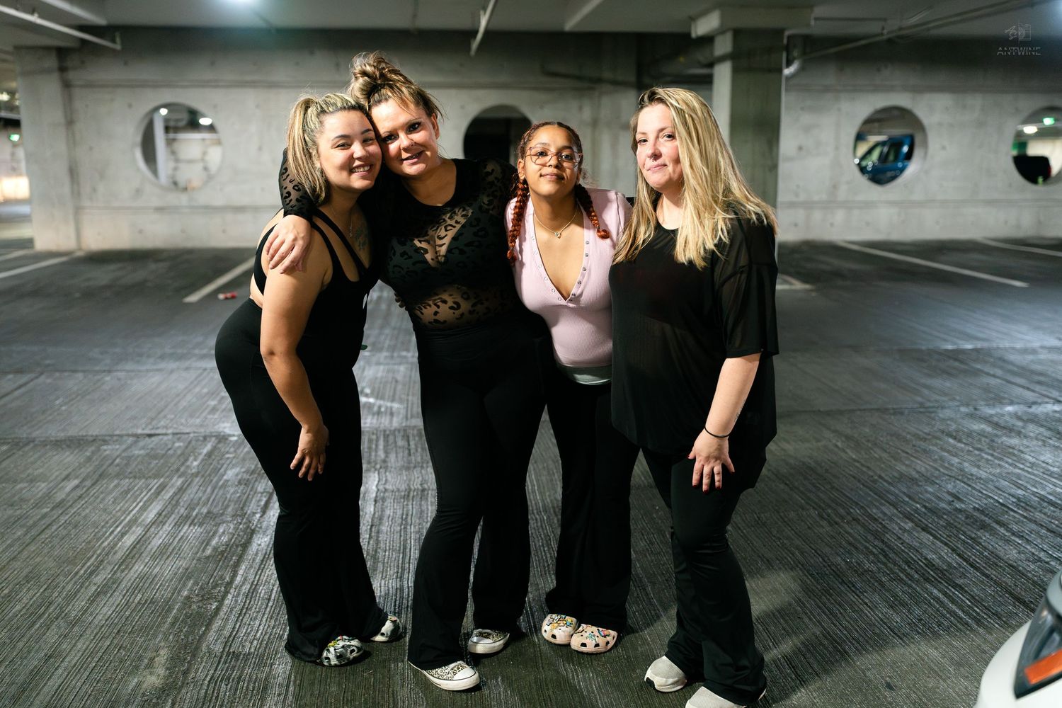 Four women in black outfits smile and pose together in a dimly lit parking garage.