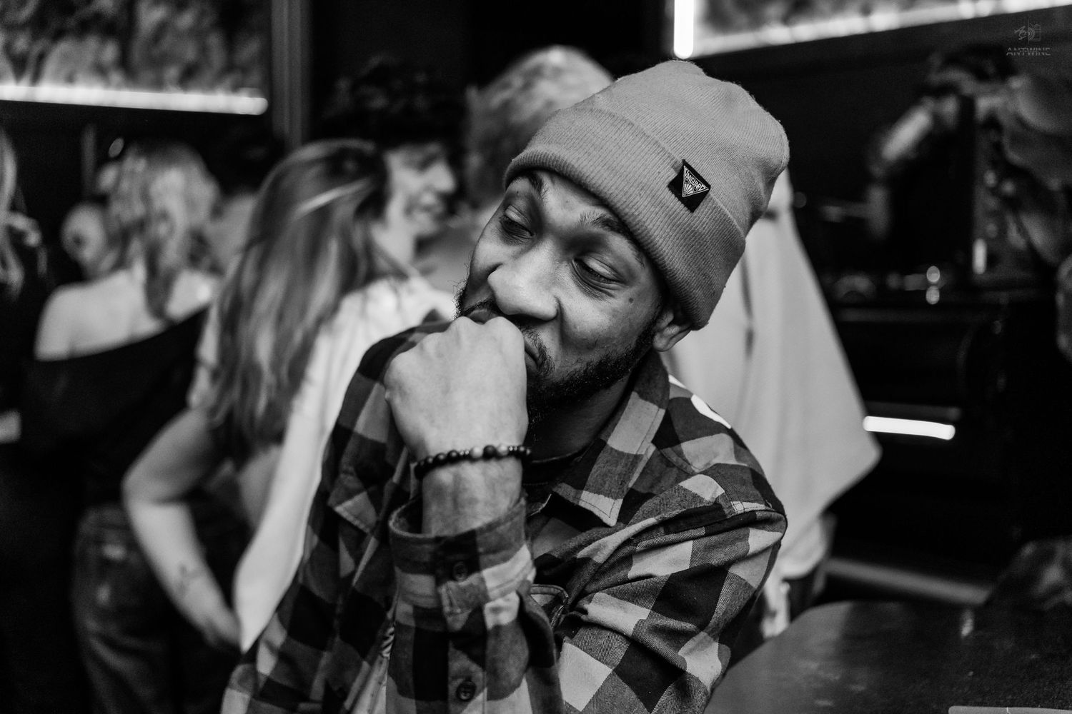 Man in plaid shirt and beanie posing thoughtfully at a nightclub party in black and white.