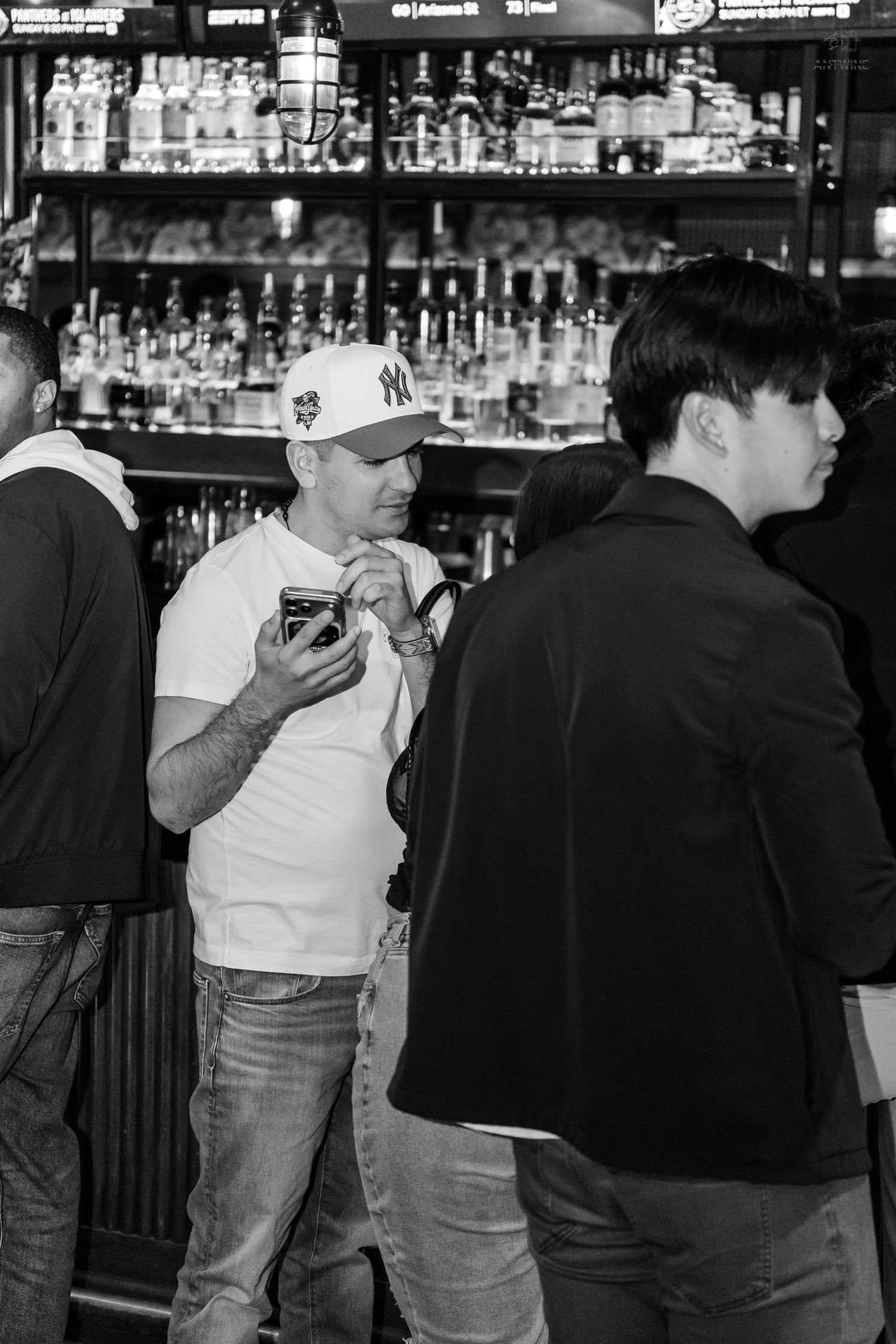 Man in white NY Yankees cap checking his phone at a crowded bar with bottle-lined shelves behind.