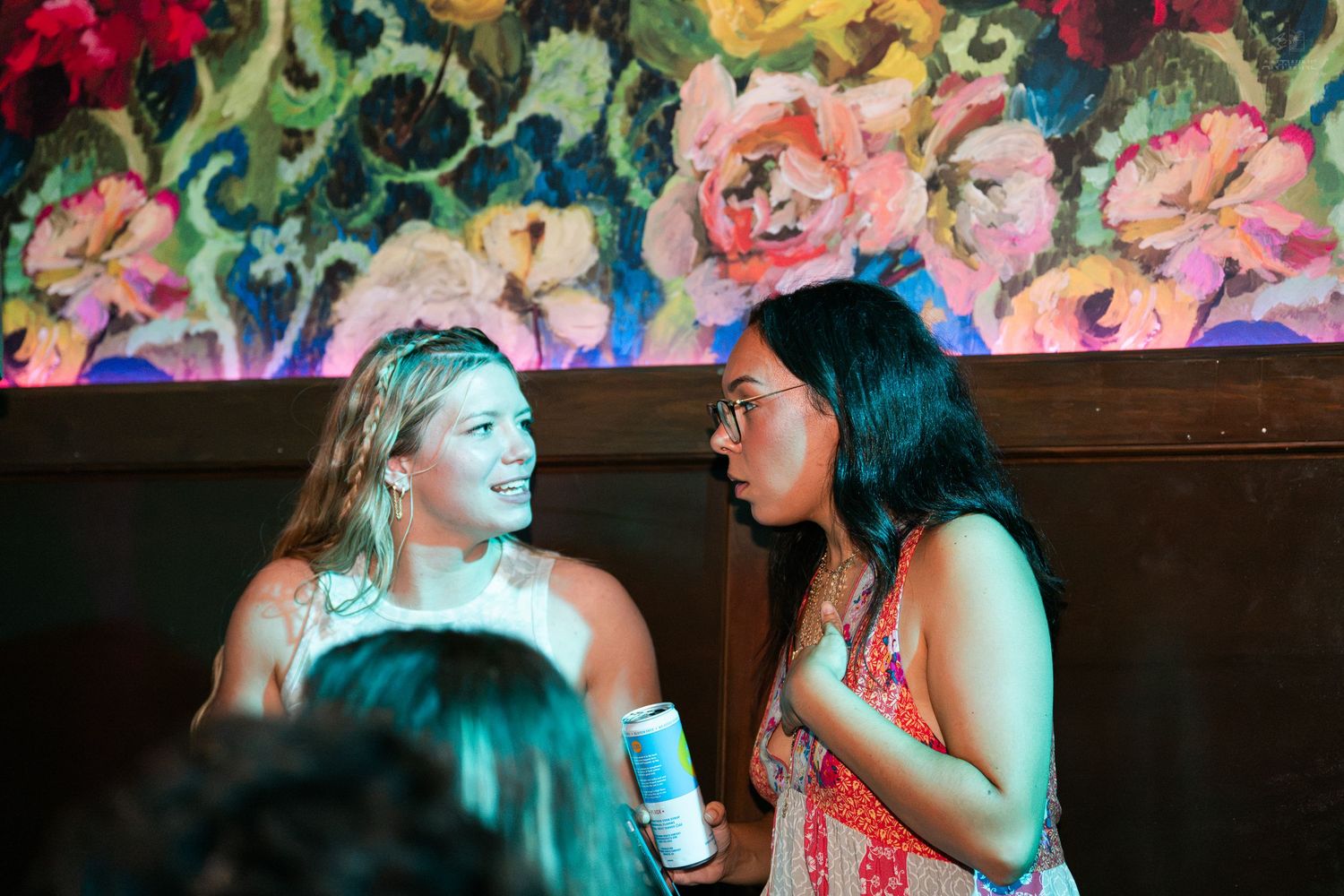 Two women laughing and chatting at a vibrant bar with colorful floral wall art.