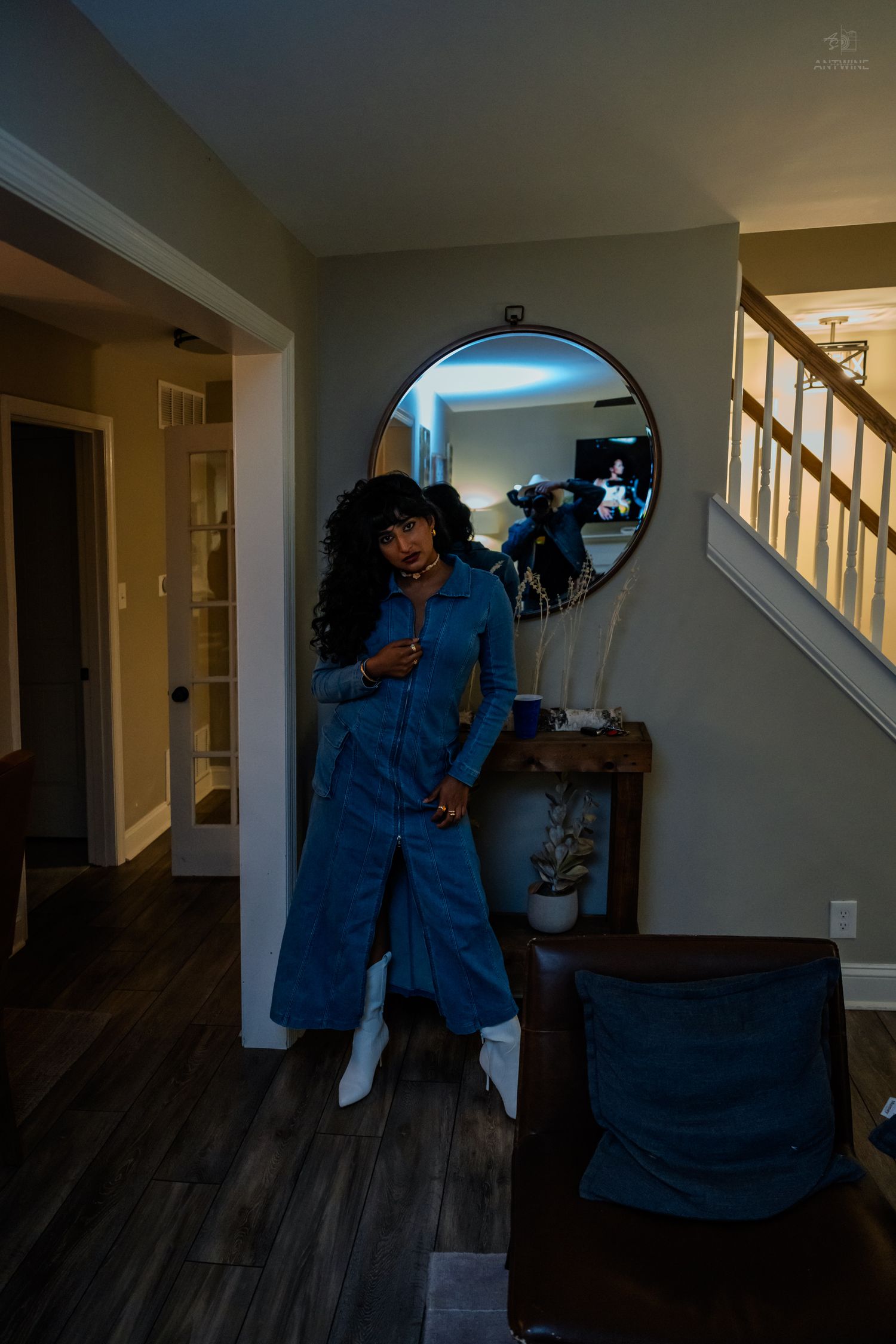 Woman in blue denim jumpsuit with white boots posing near round mirror in home entryway at night.