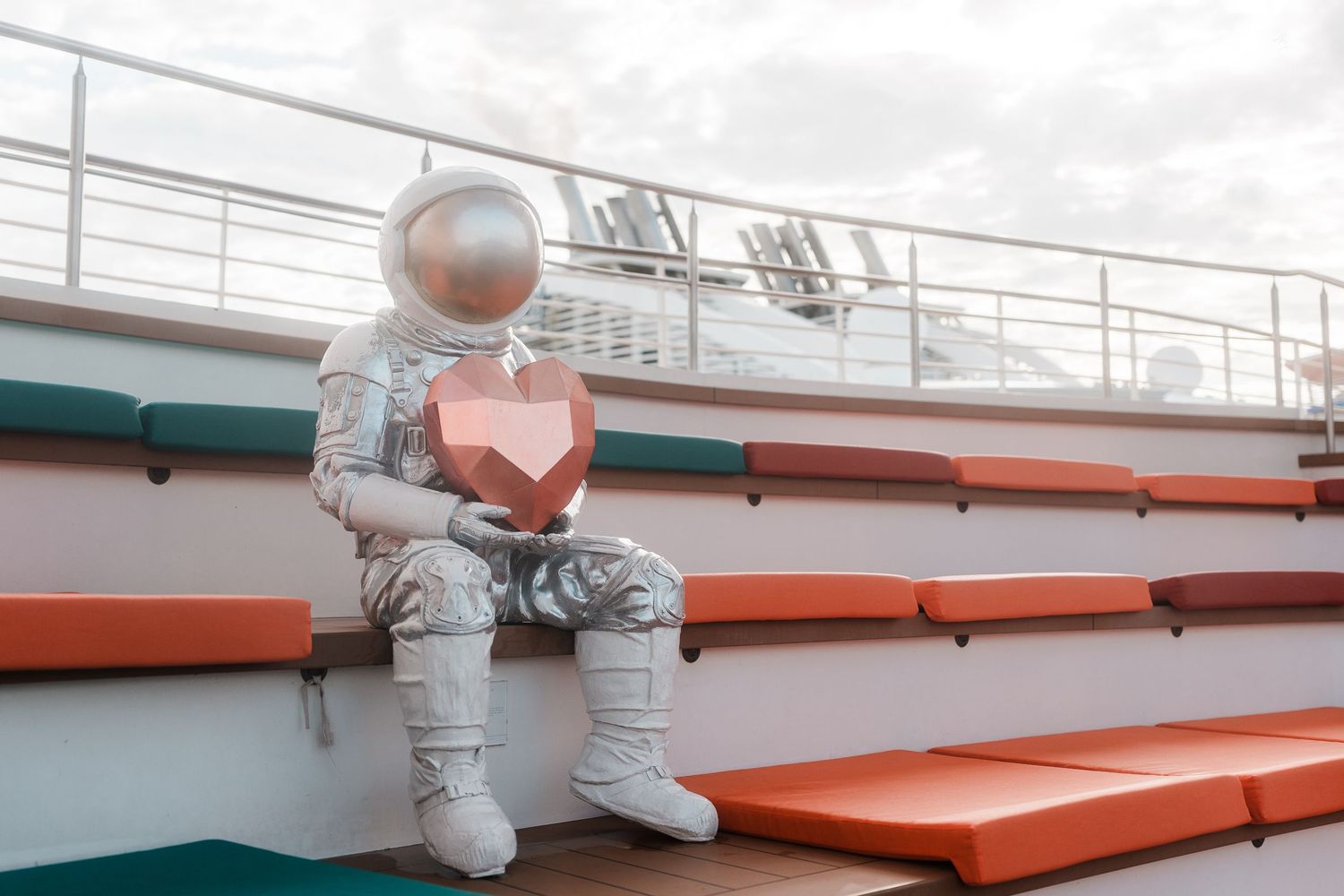 An astronaut statue holding a geometric heart sits on colorful bleachers on a cruise ship deck.