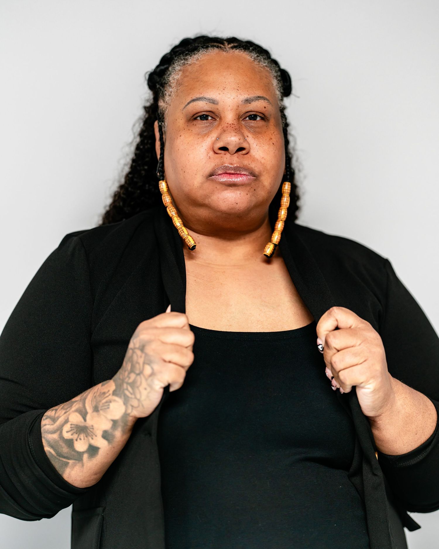 Black woman in black outfit gripping lapels, showing floral arm tattoo and yellow hoop earrings.