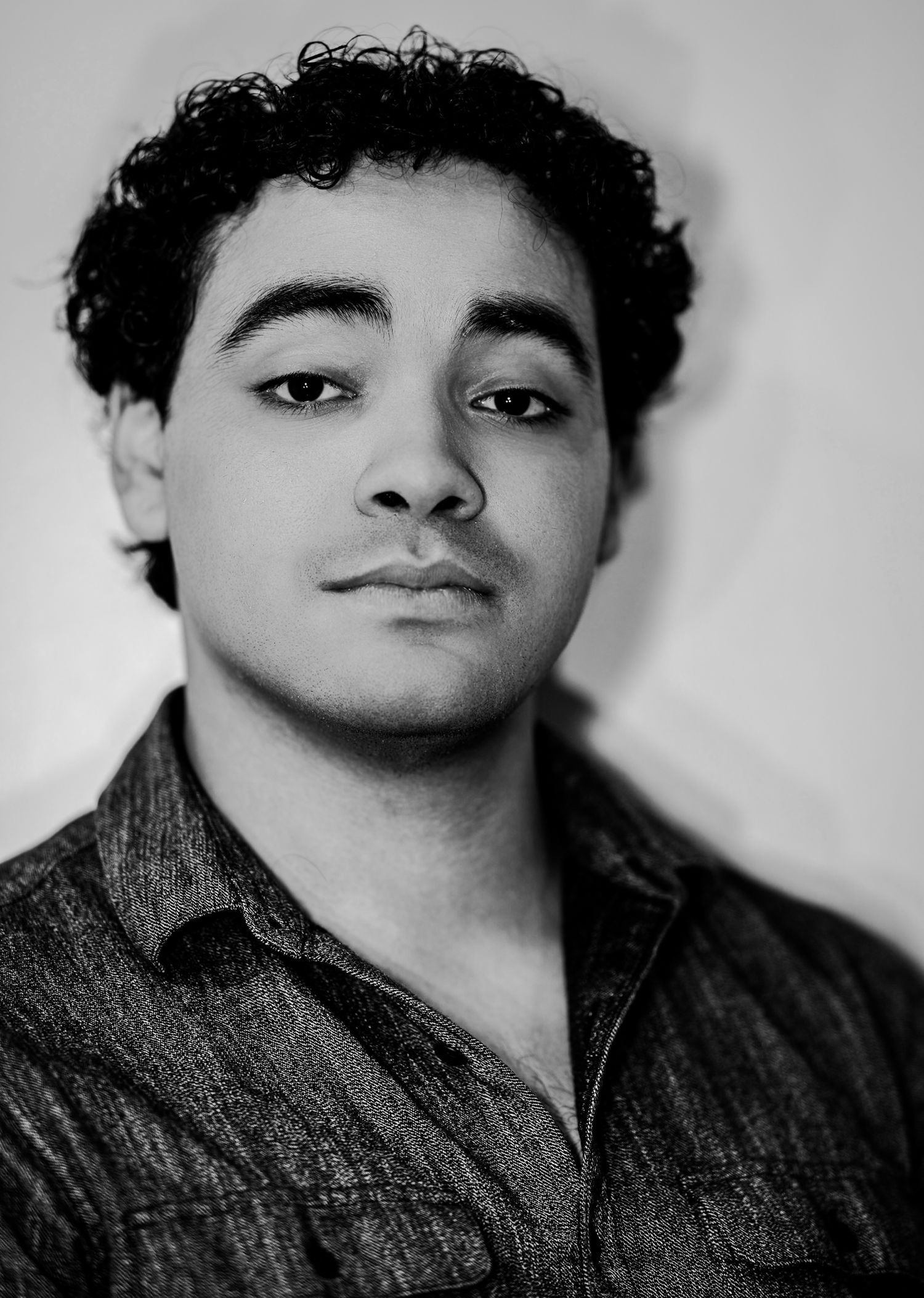 Young man with curly dark hair in a dark shirt gazing forward in a dramatic black and white portrait.